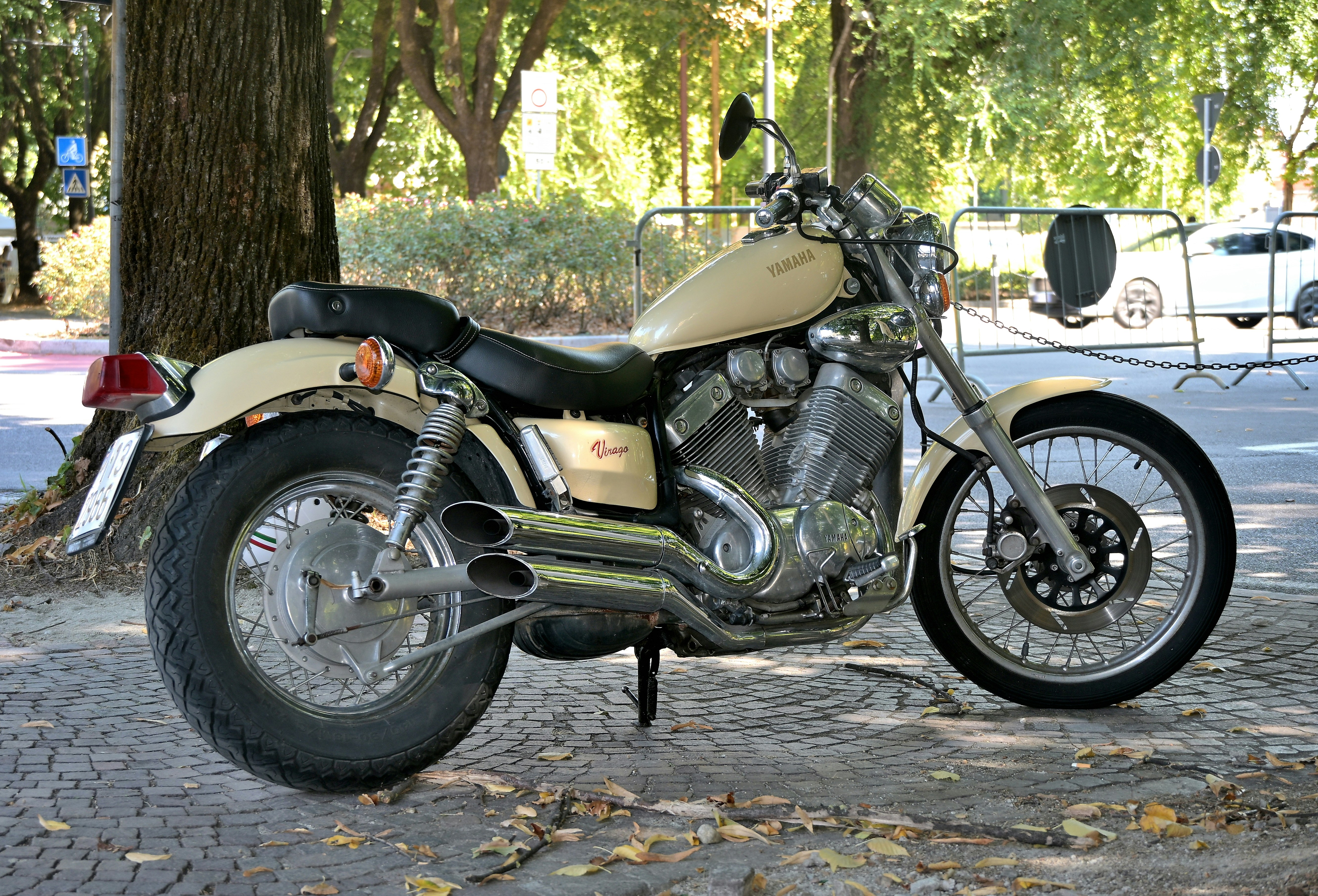 Uma motocicleta Yamaha Virago cor creme estacionada em um caminho de paralelepípedo perto de uma árvore