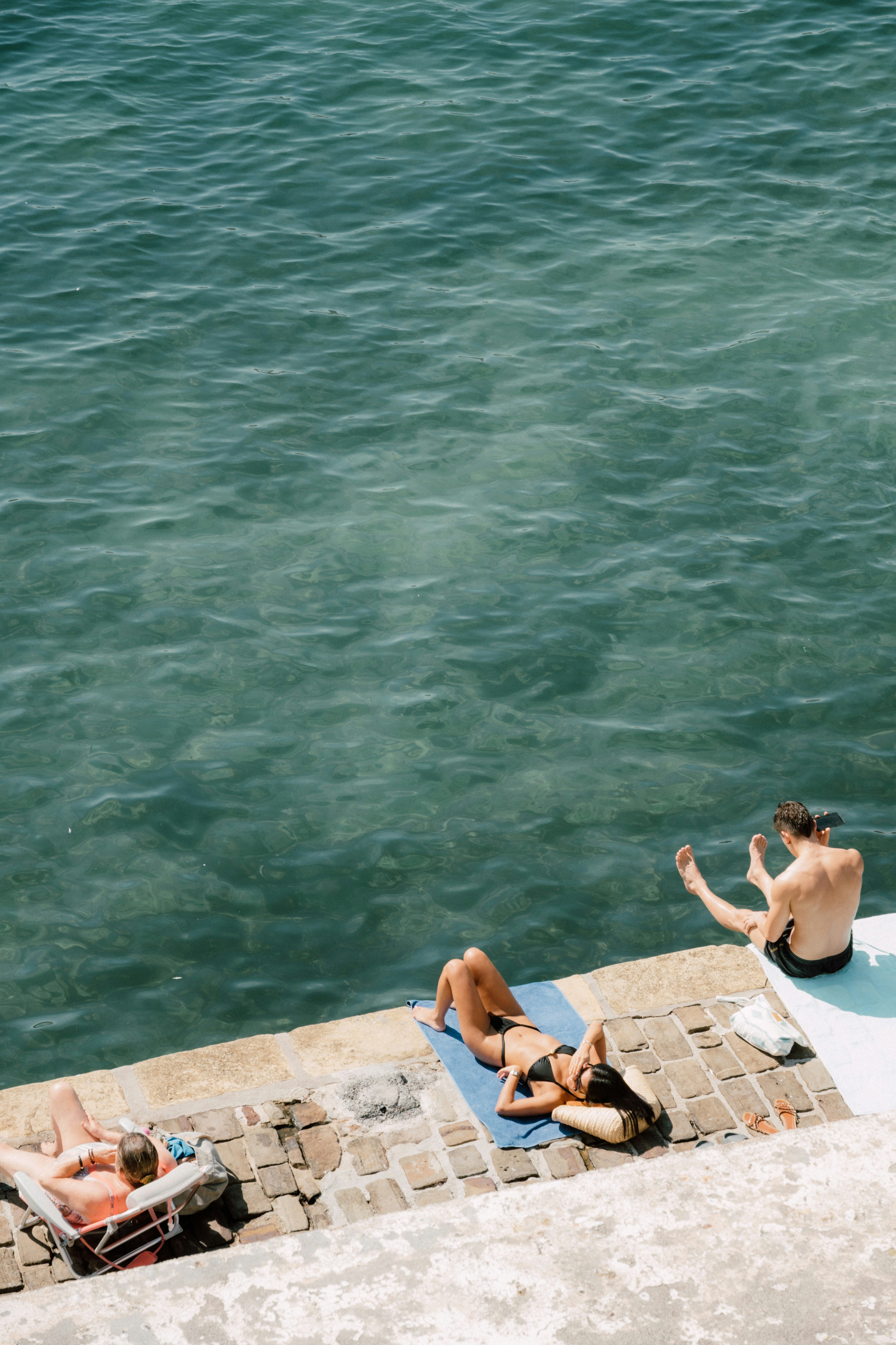 Three people relaxing on a stone quay beside clear green sea water