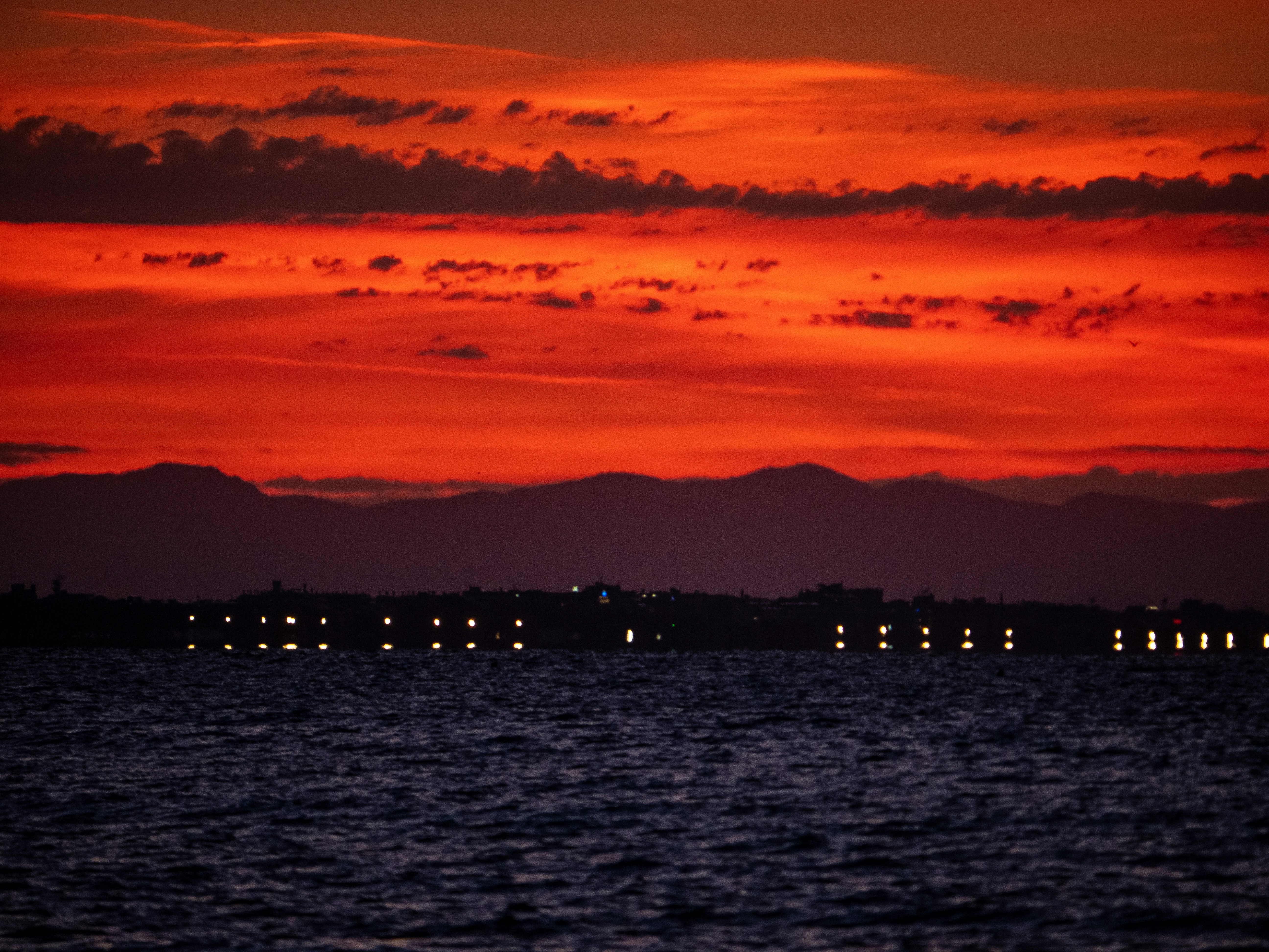 A dark city skyline with glowing lights across water under a vibrant red sky