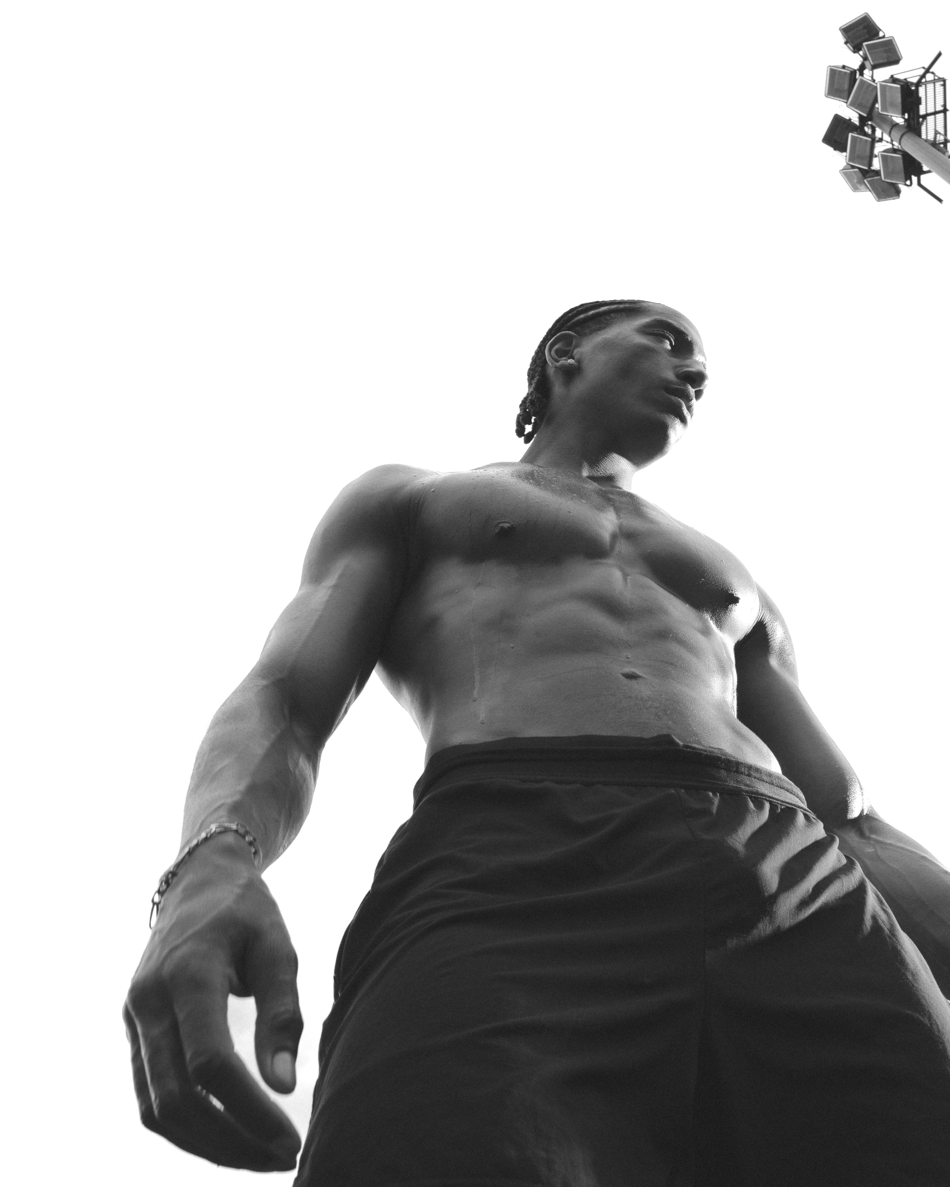 A shirtless man with braided hair in black shorts under stadium lights