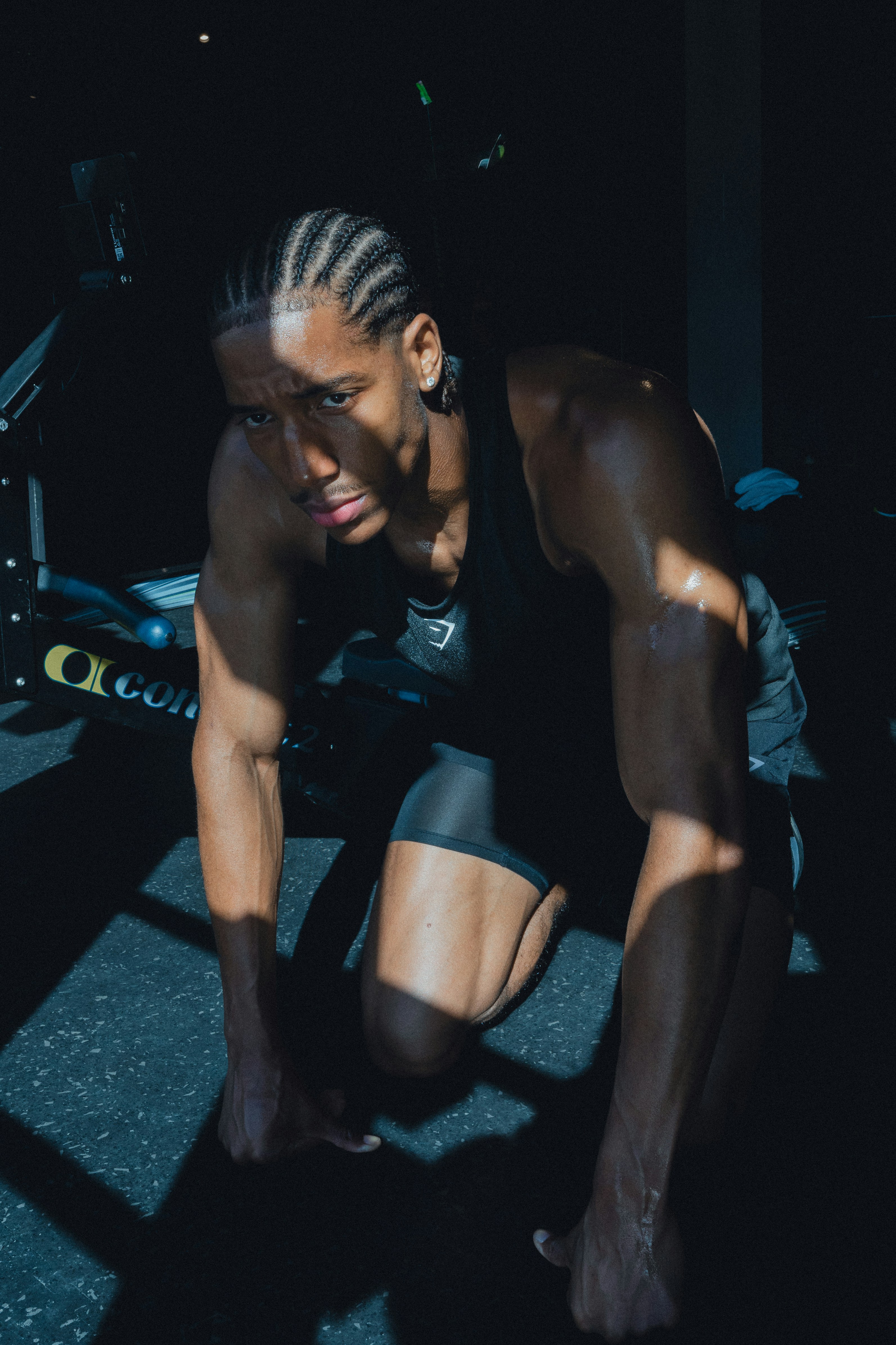 A man with braided hair kneeling on a gym floor in low light