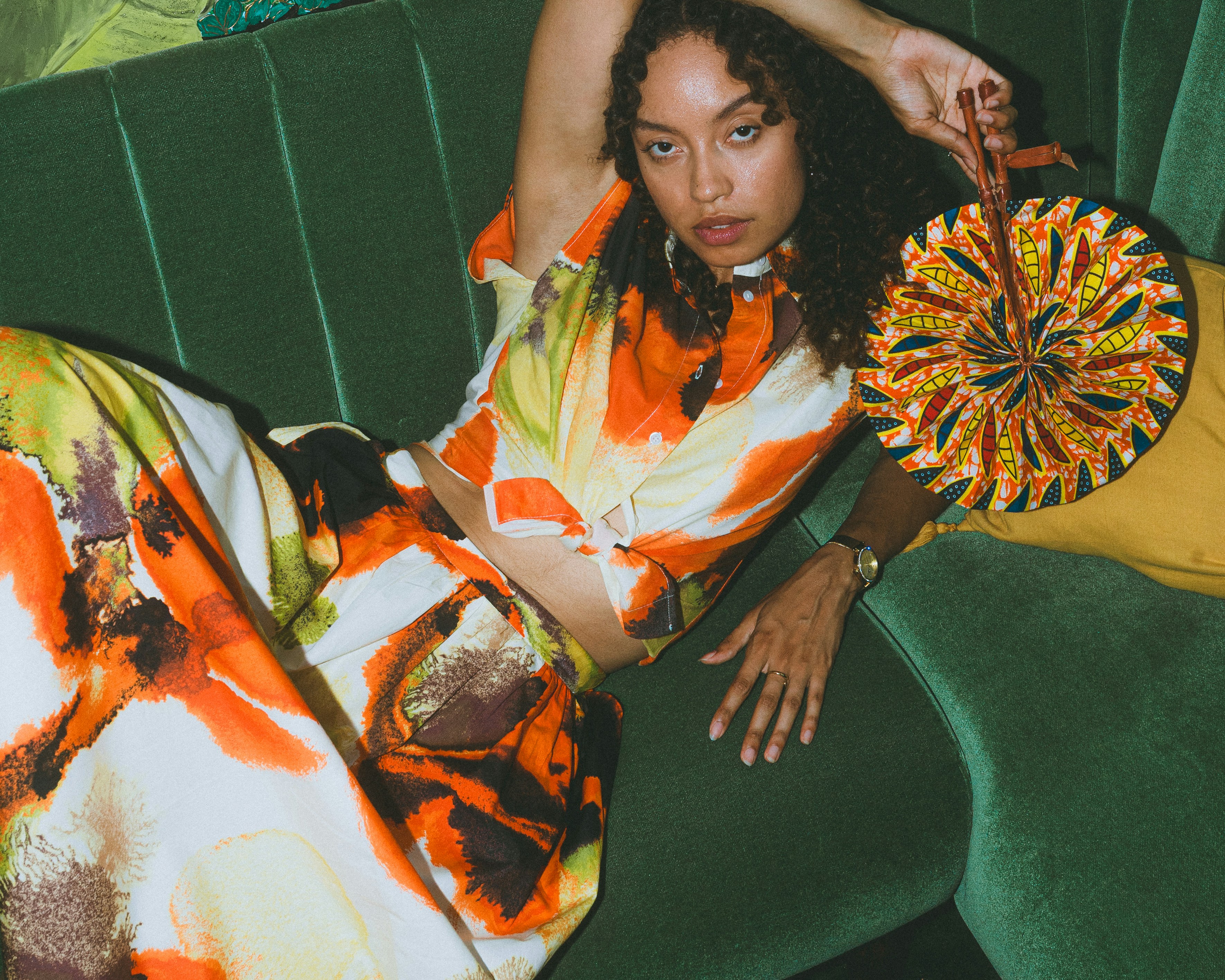 Woman in a colourful patterned outfit holding a round fan on a green sofa