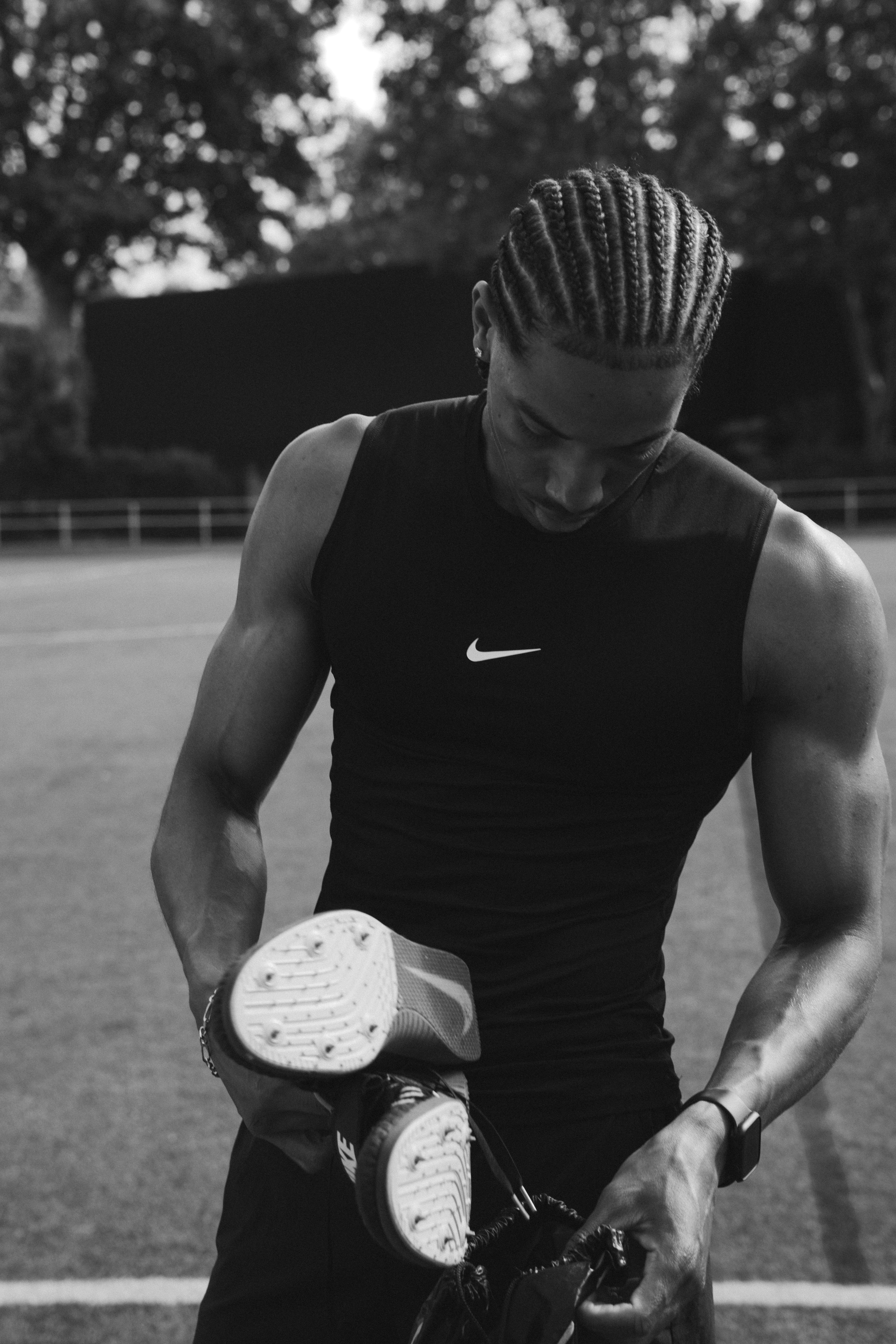 A man with braided hair holding track spikes on an outdoor athletic field