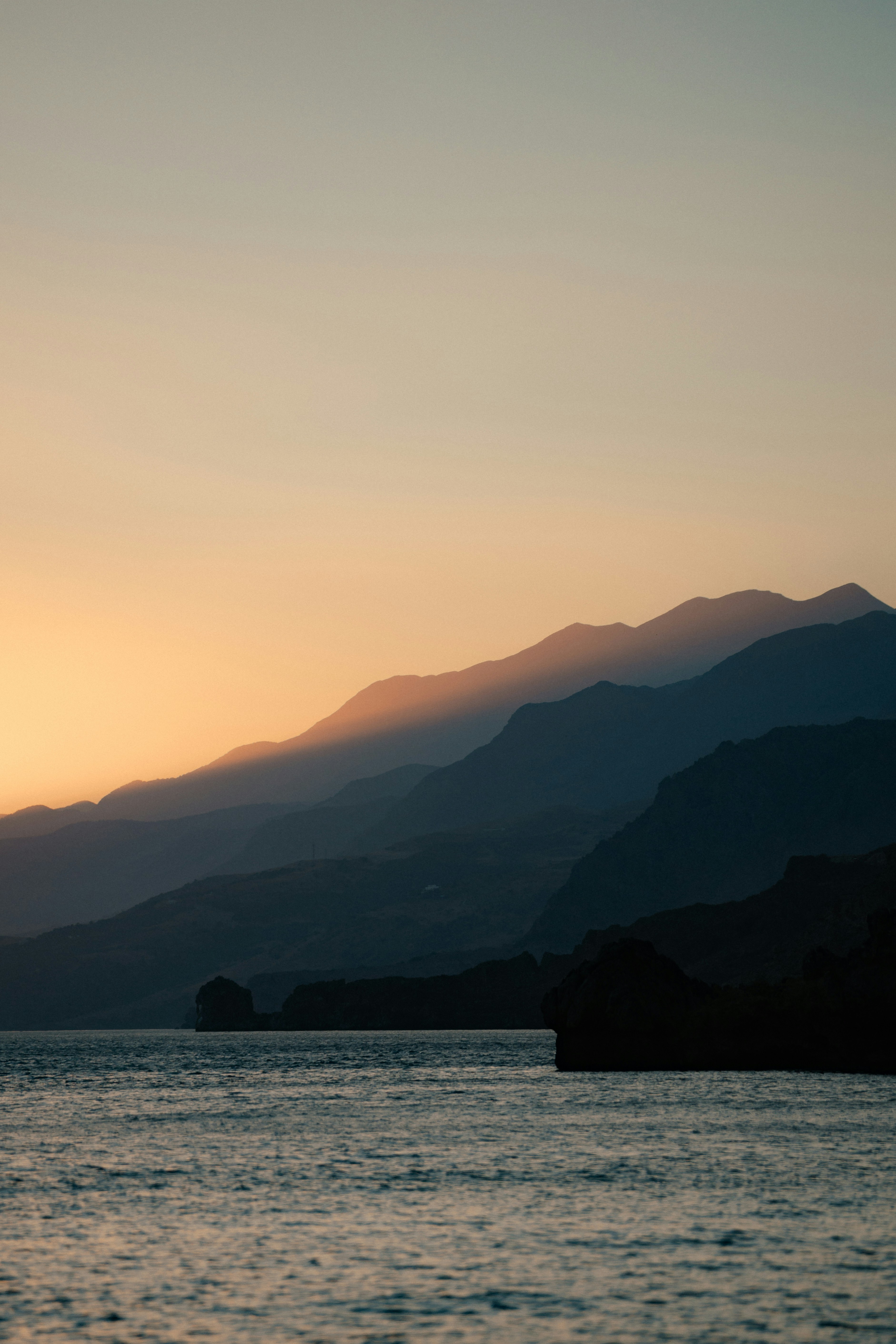 Silhouetted mountains rising above a calm sea under a warm orange sky