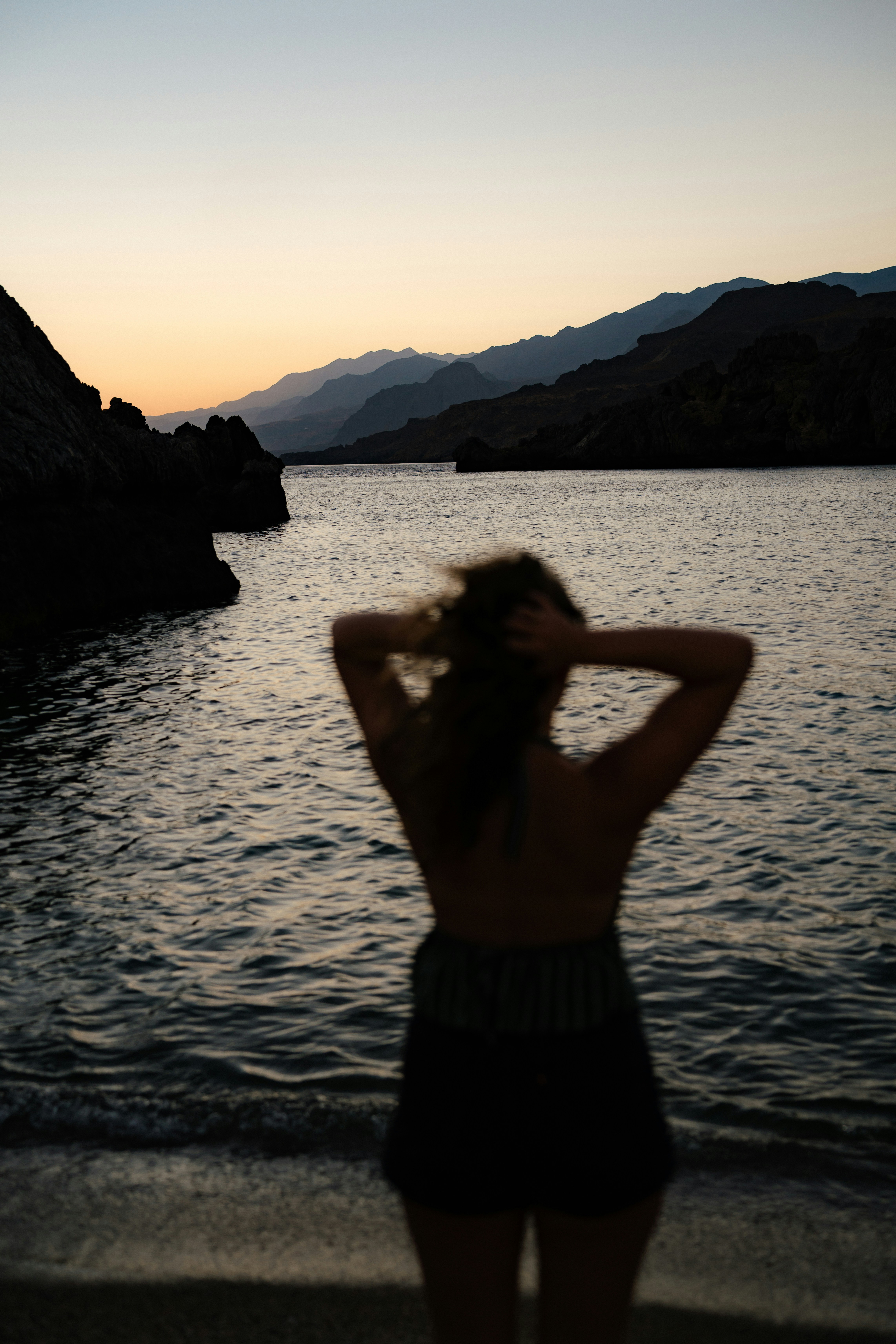 A person in silhouette with hands in hair facing a calm sea and mountains