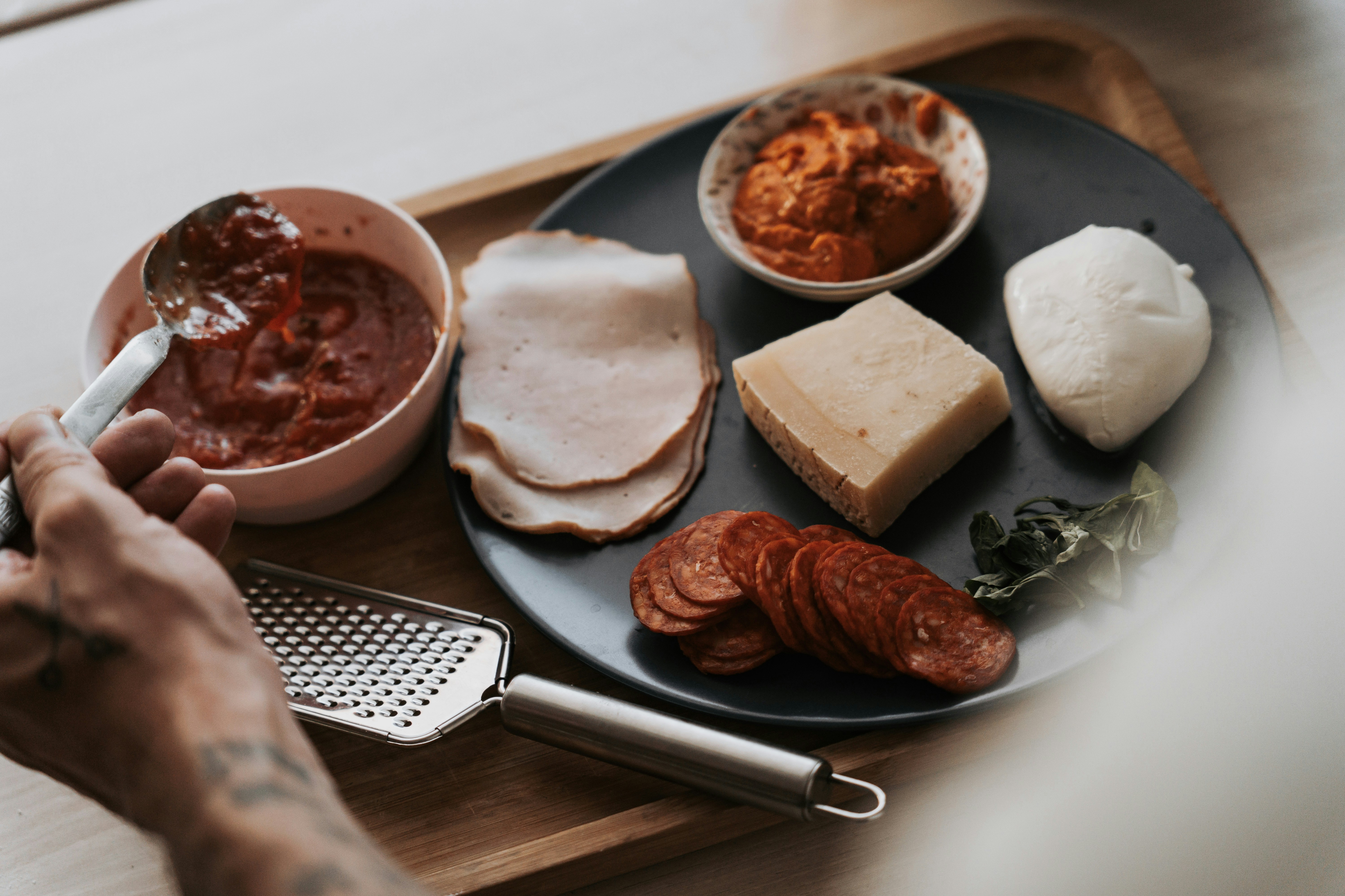 A hand holding a spoon over tomato sauce with deli meats and cheeses
