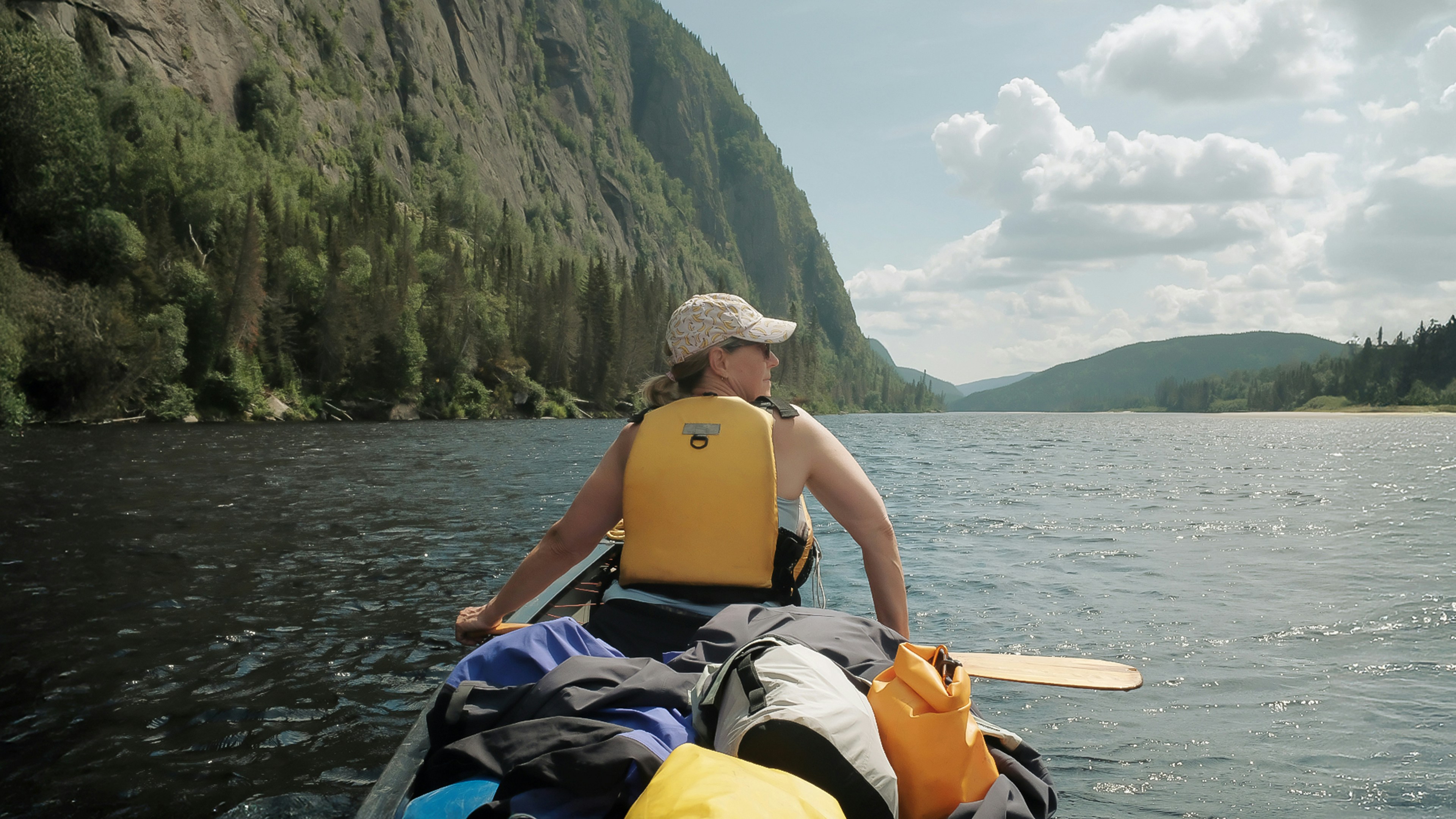 Paddler in a yellow life jacket steering a loaded canoe, a yellow roll-top dry bag and gear packed in front of her, a steep forested shore ahead