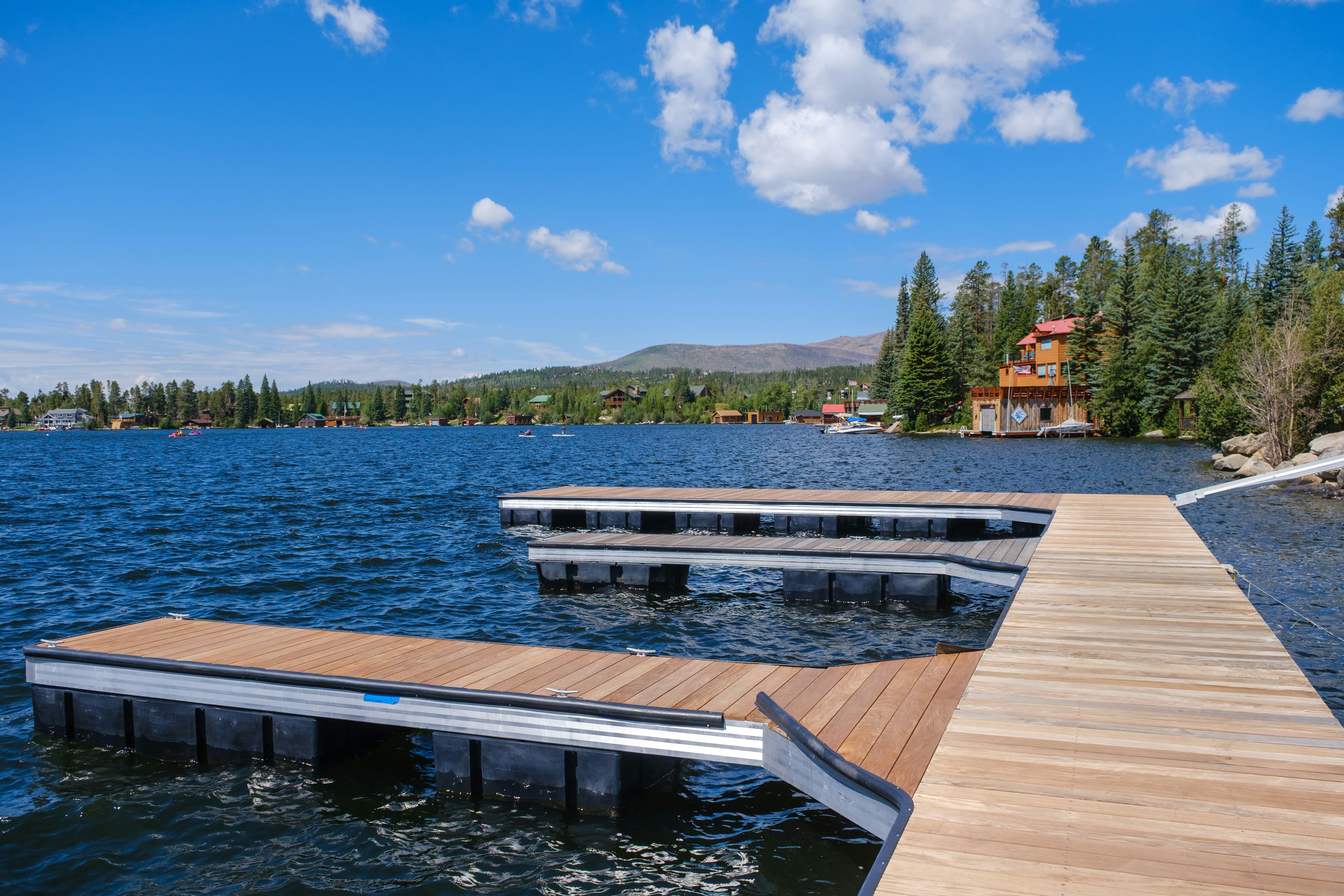 Sectional wooden floating dock reaching into a deep blue lake, with pine trees and lakefront cabins along the far shore