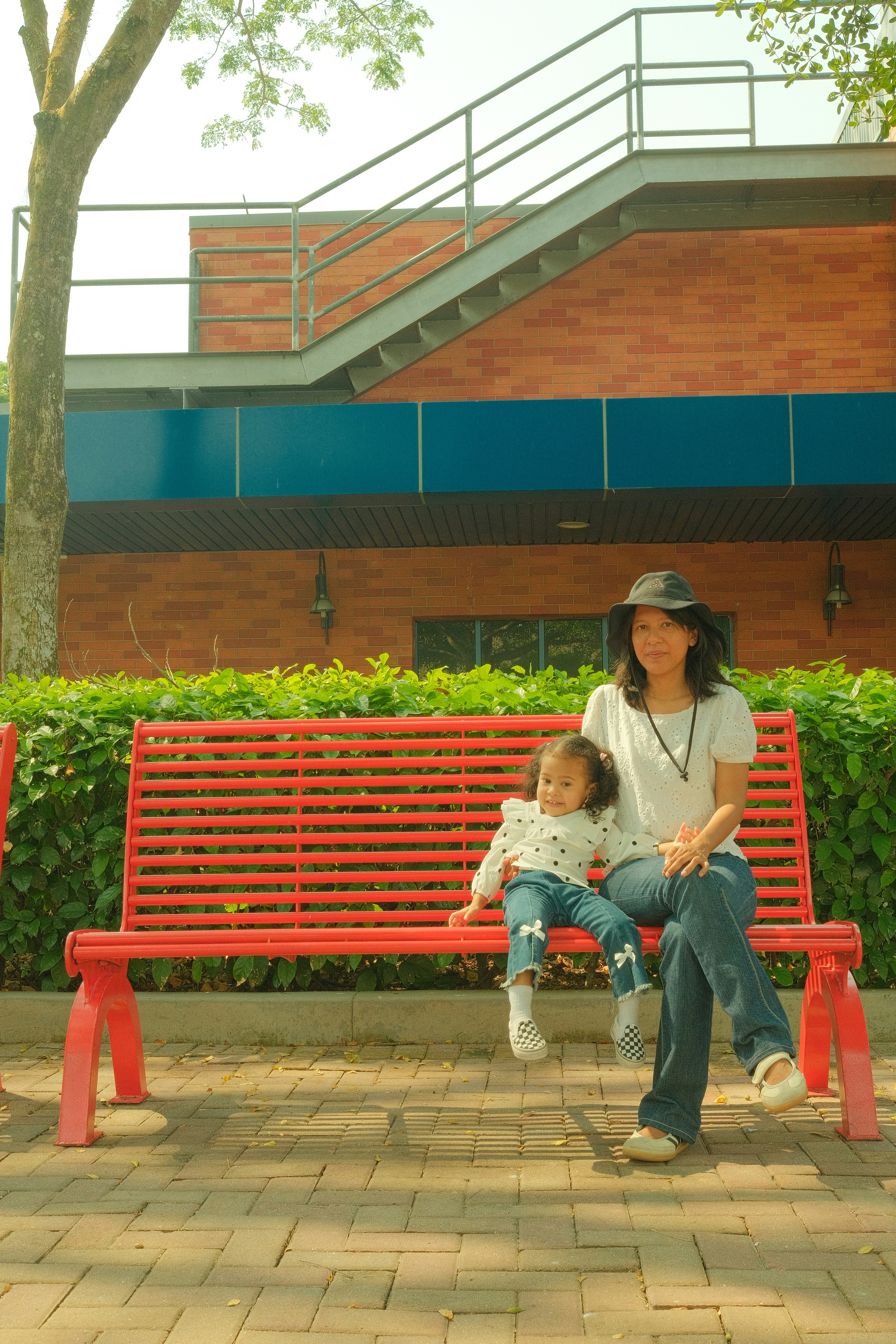 Une femme et un enfant sur un banc rouge devant un bâtiment en briques