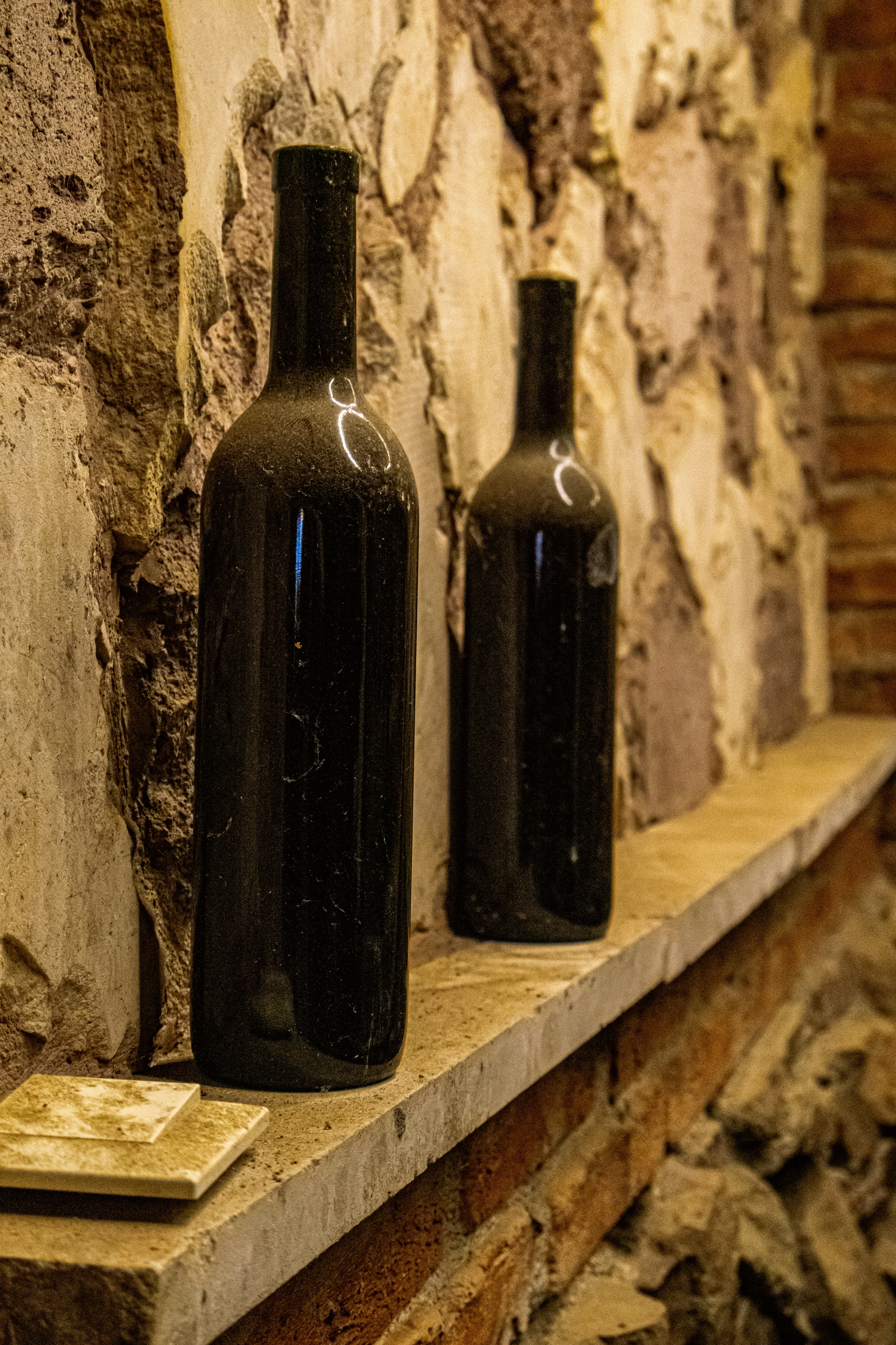 Two dark glass wine bottles on a stone ledge against a rustic wall