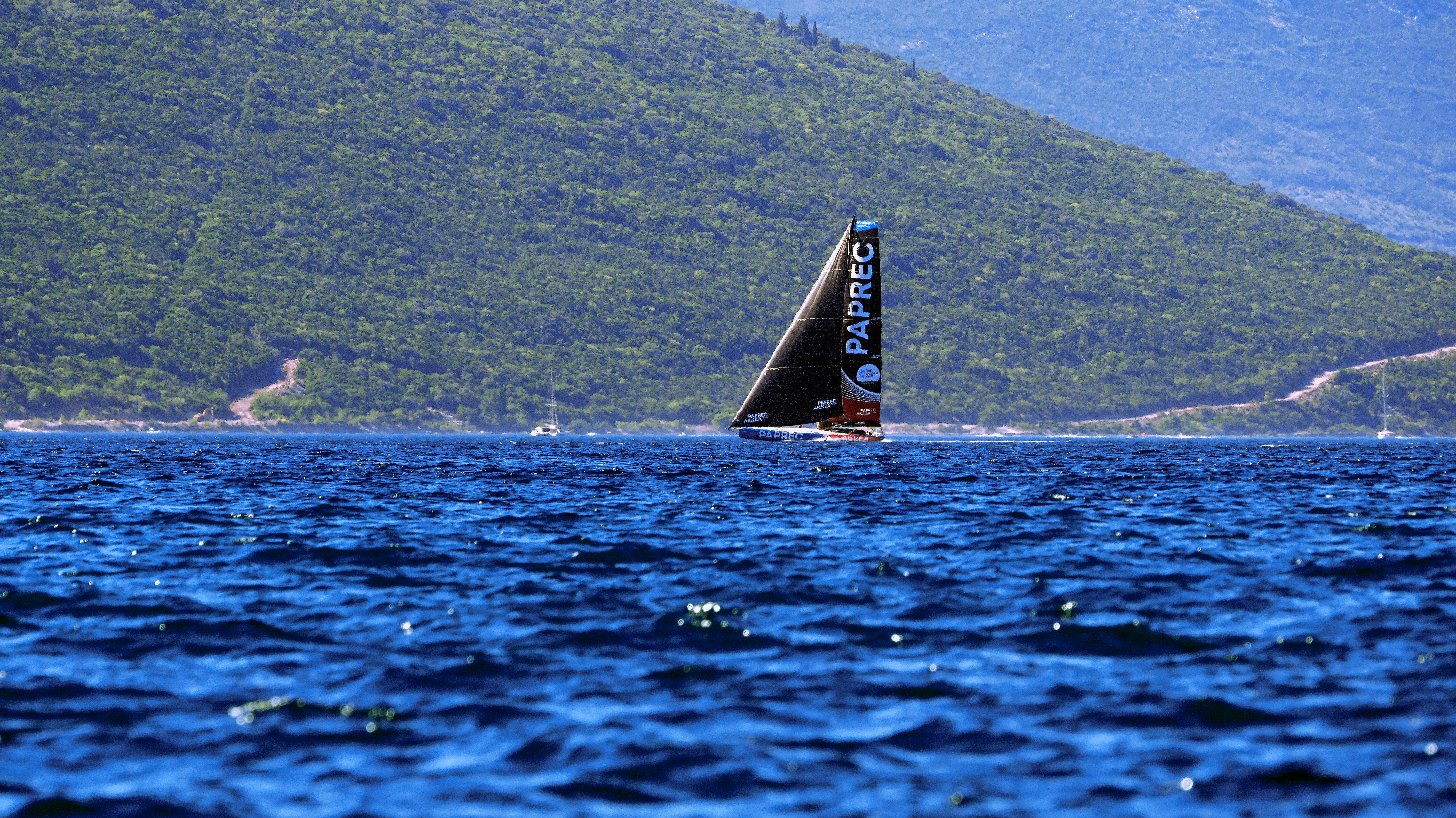 A racing sailboat with a black sail on blue water before a green mountain