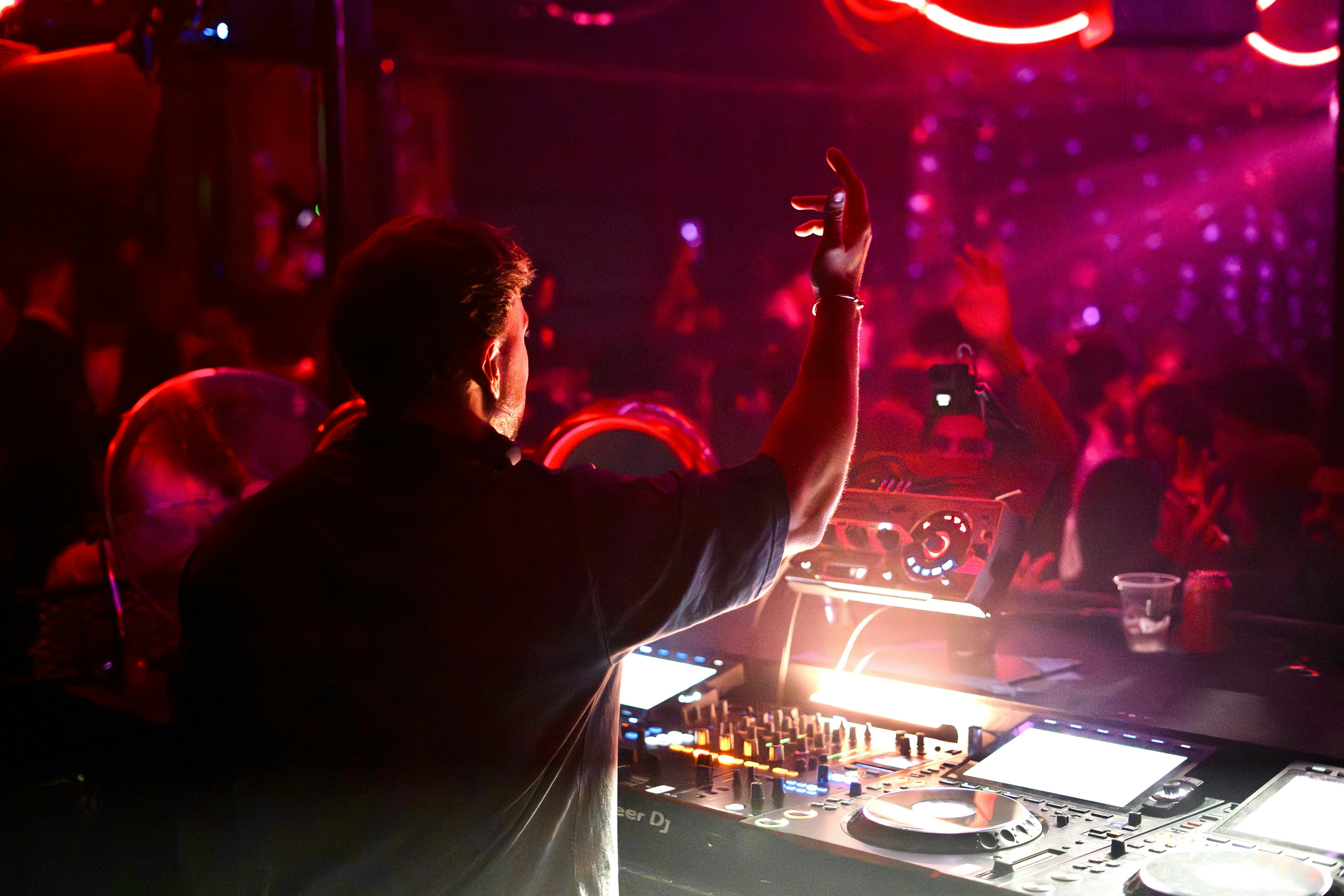 A DJ with one arm raised at a mixing console in a dark nightclub