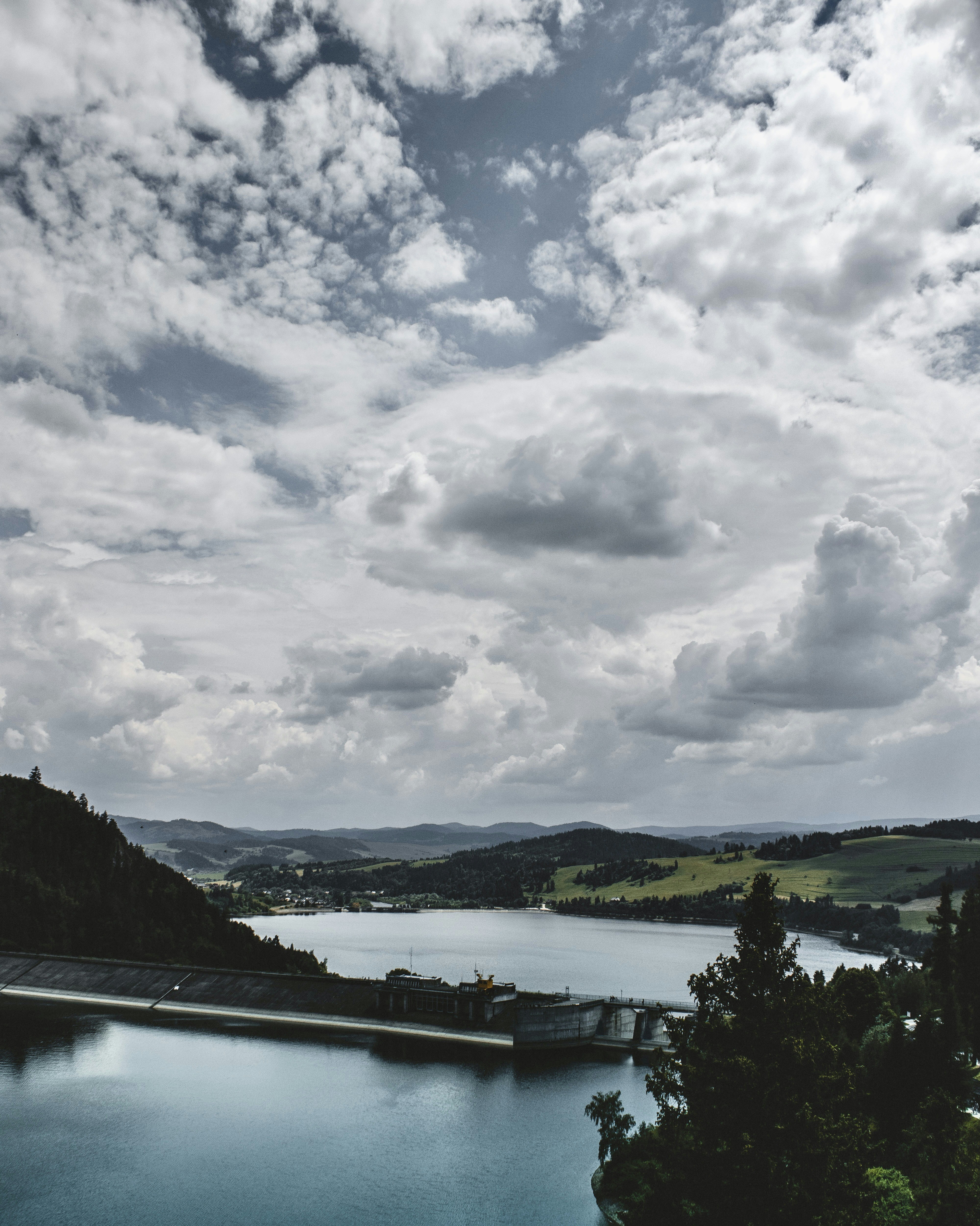 A concrete dam on a large lake surrounded by green hills under cloudy skies