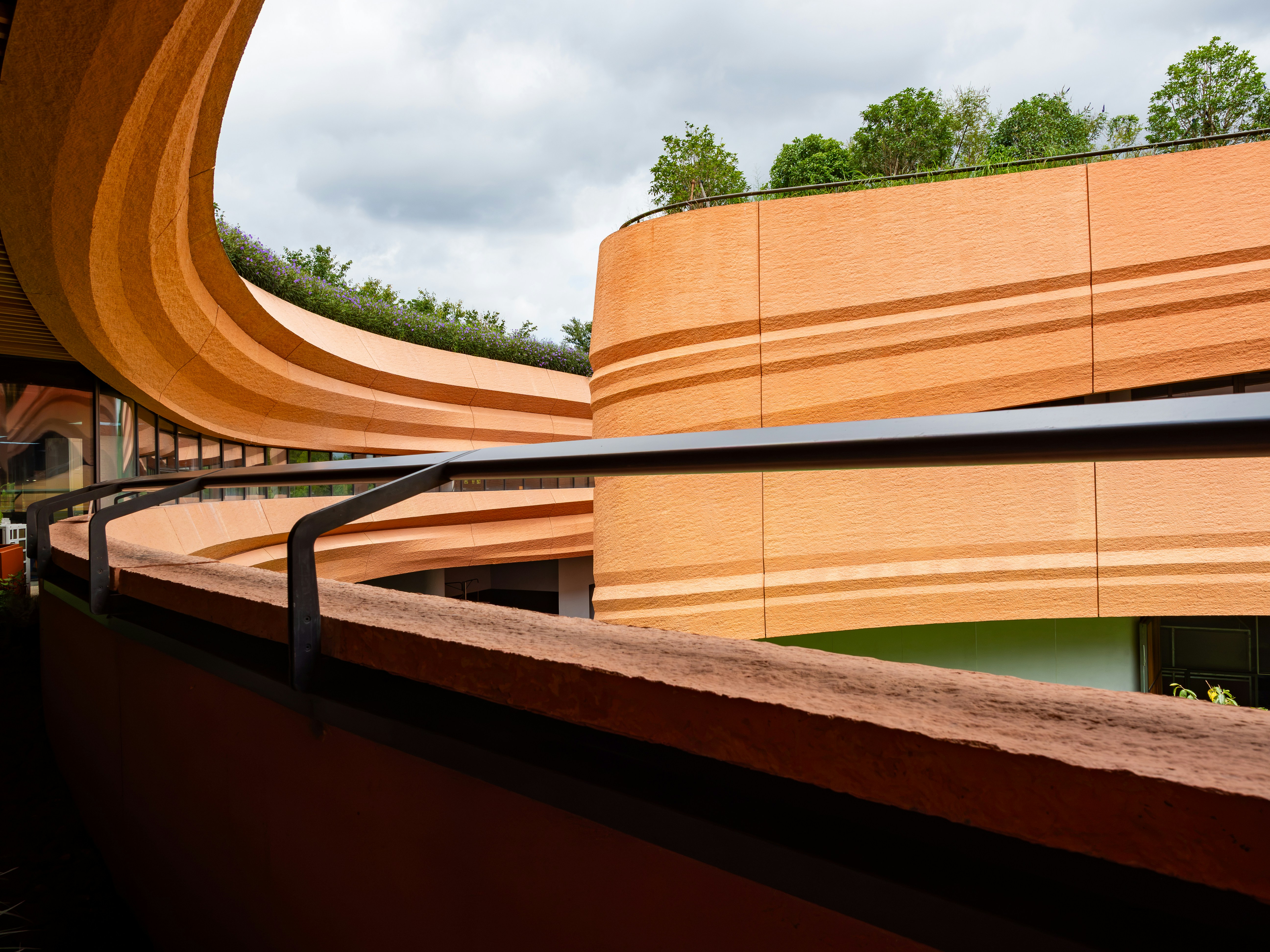 Curved orange walls of a modern building with a rooftop garden under cloudy skies