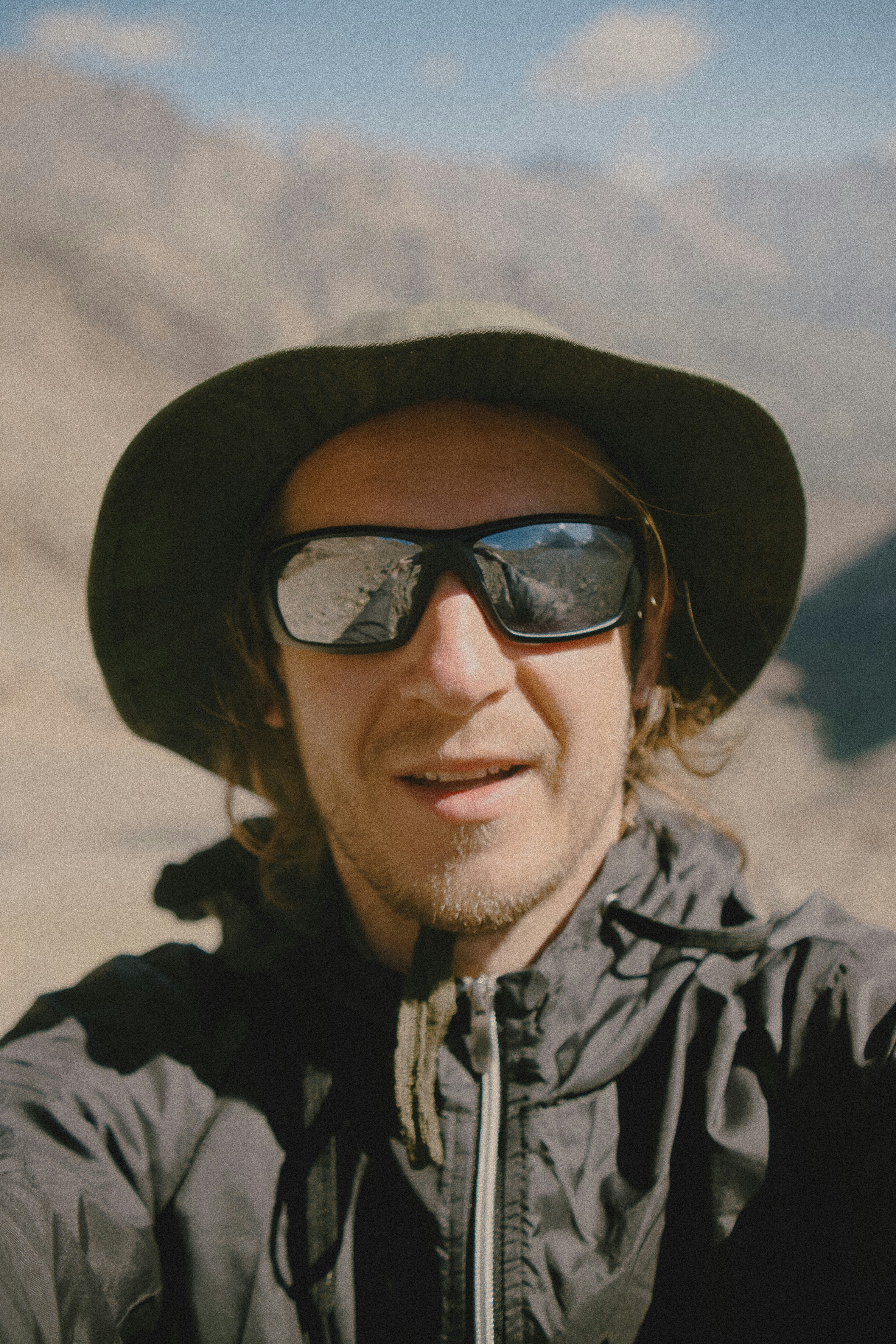 Ein Mann mit Bucket Hat und Sonnenbrille vor einer verschwommenen Berglandschaft