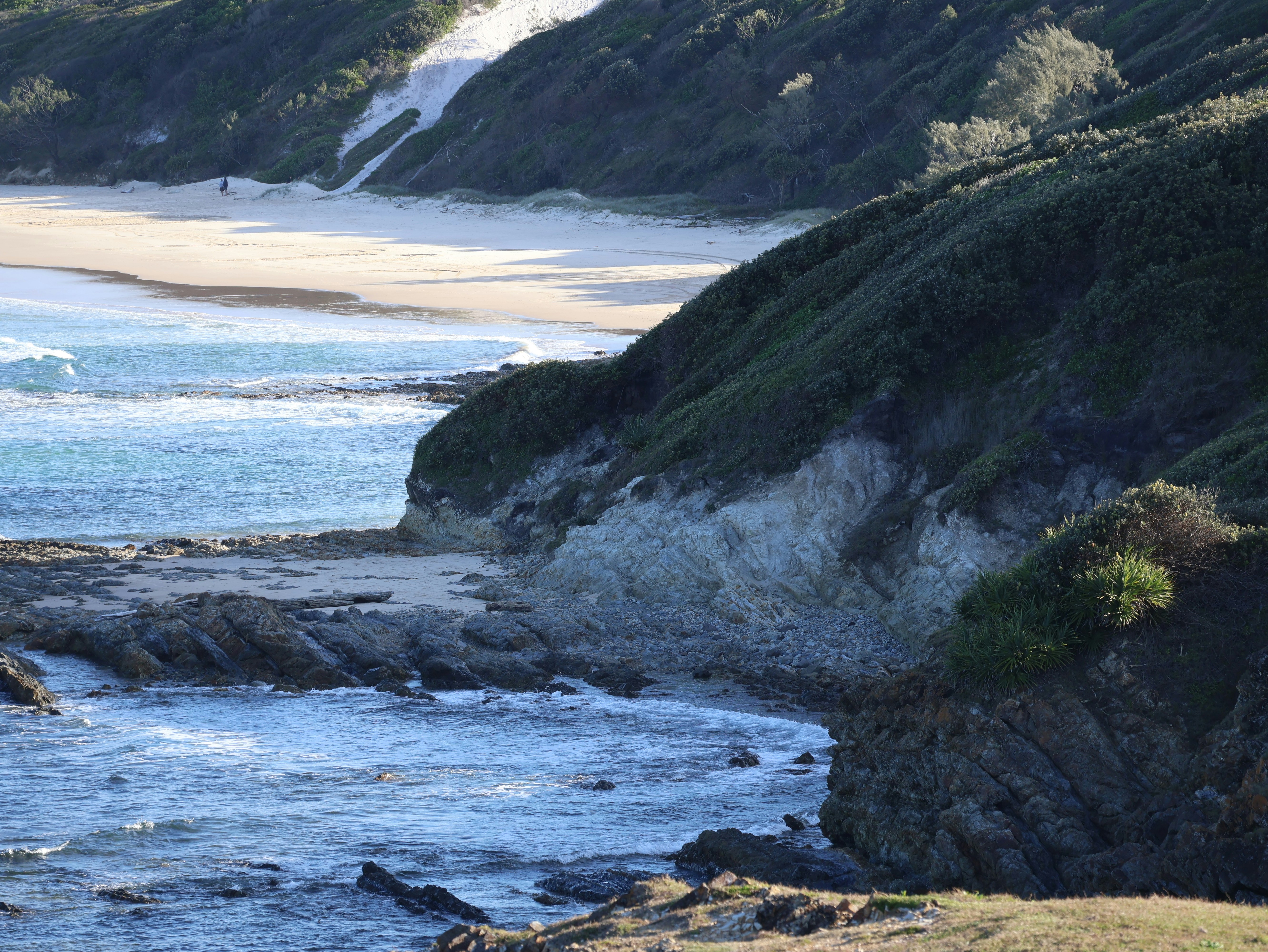 A sandy beach below a steep vegetated cliff with rocky shoreline and waves