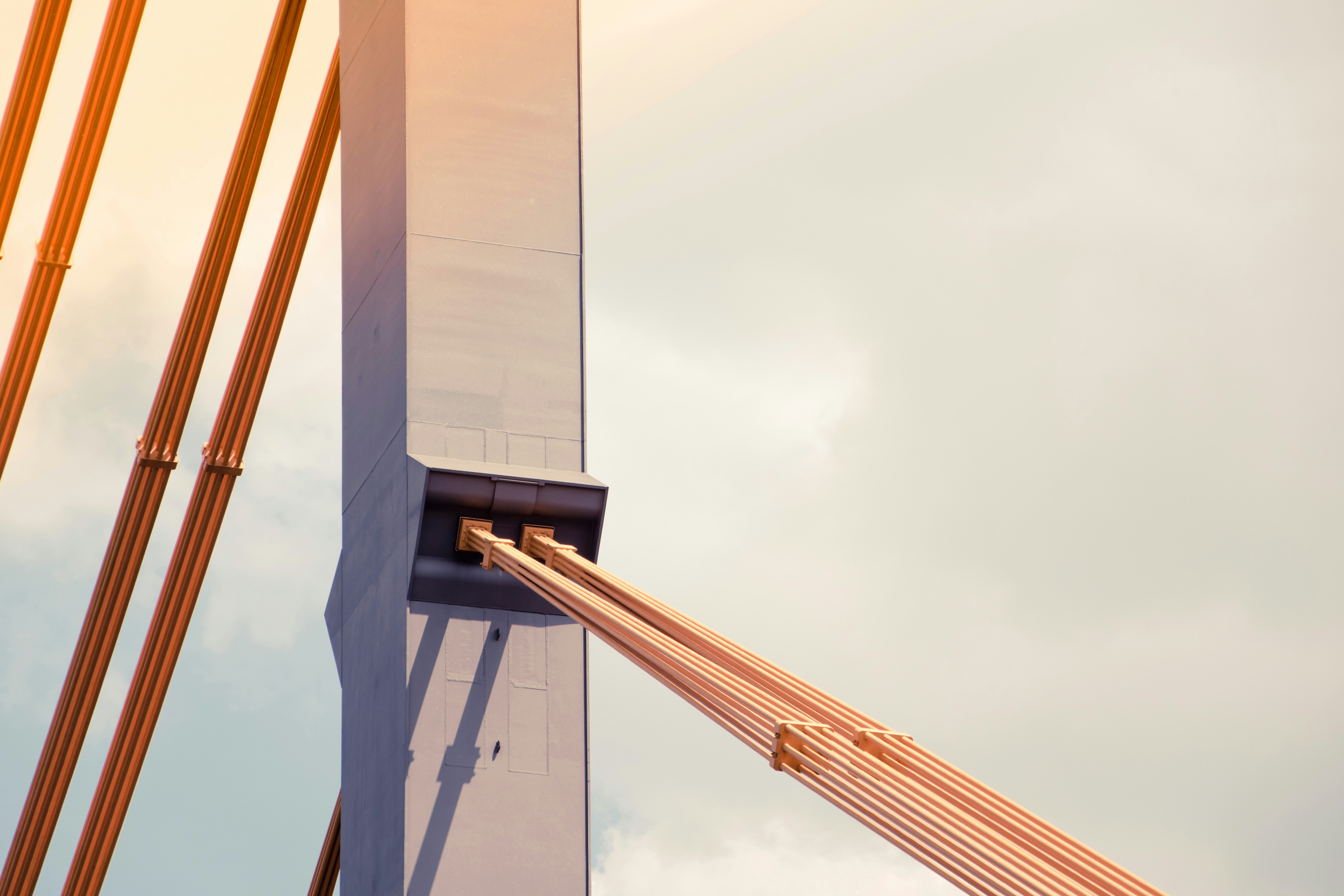 Ein grauer Betonbrückenmast mit orangefarbenem Kabel hält vor einem blassen Himmel
