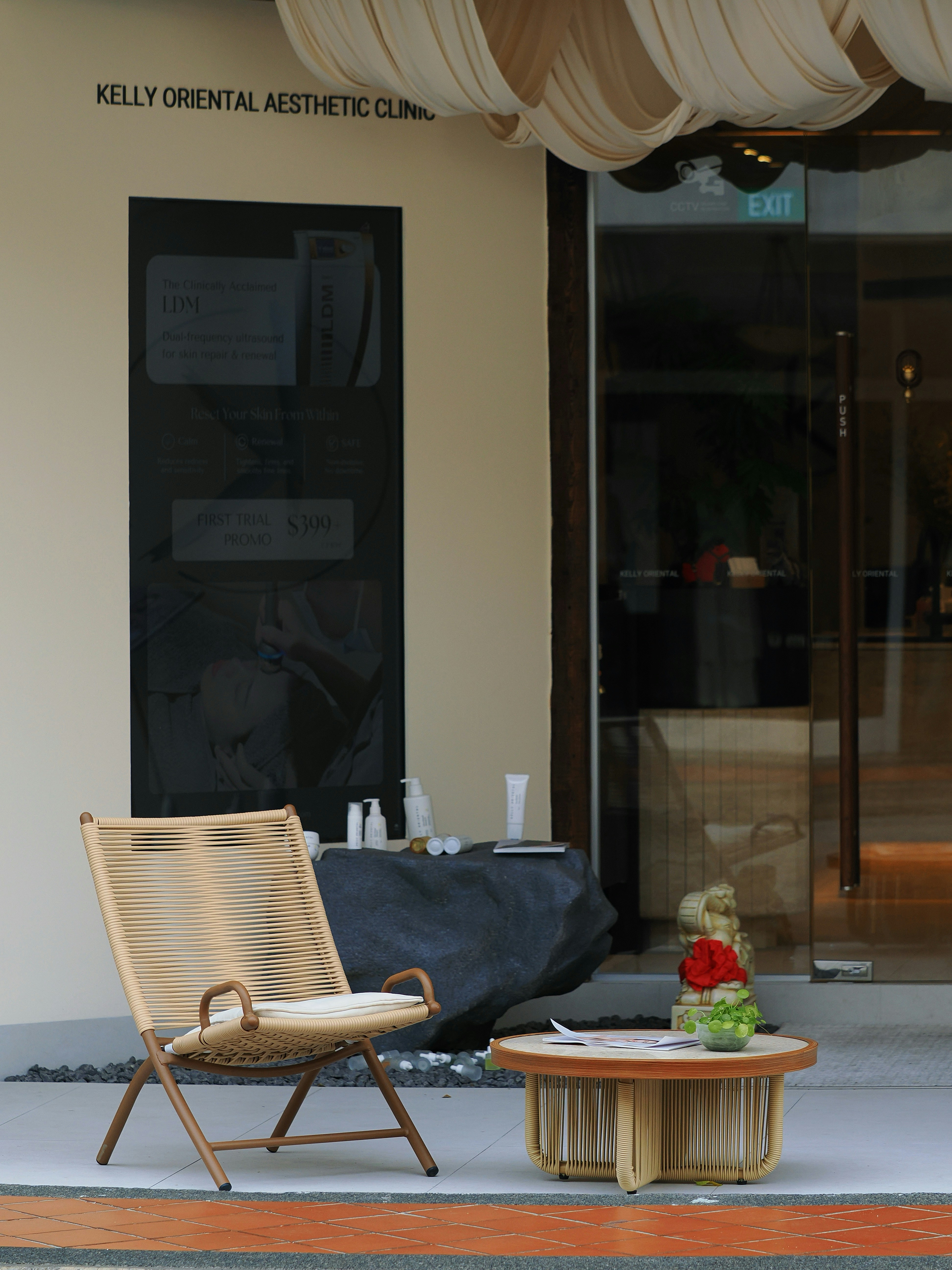 Entrada de un centro de estética: pared color crema, puertas de vidrio, un sillón de ratán y una mesita con flores sobre la vereda