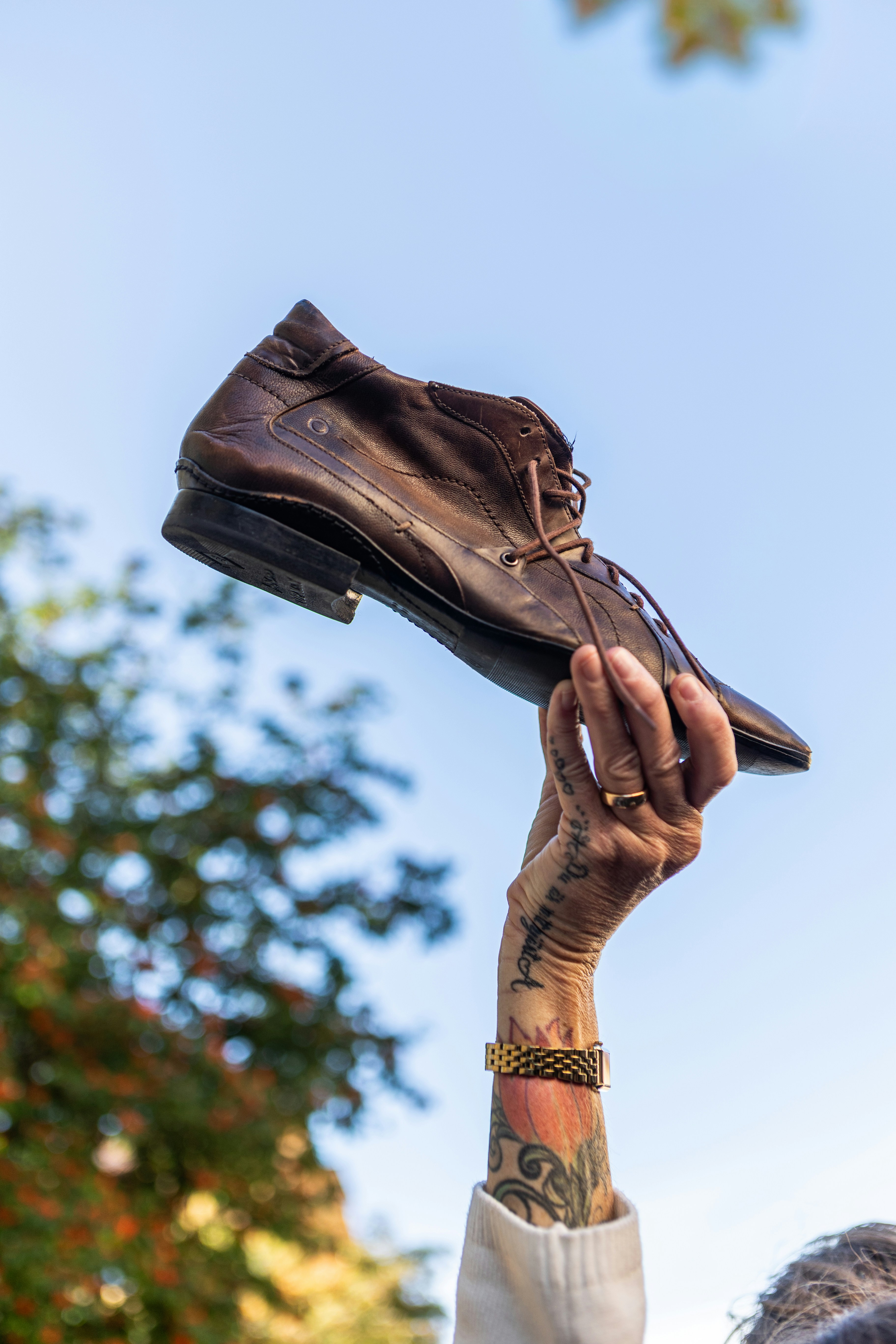 A hand with tattoos holding a brown leather dress shoe against a blue sky