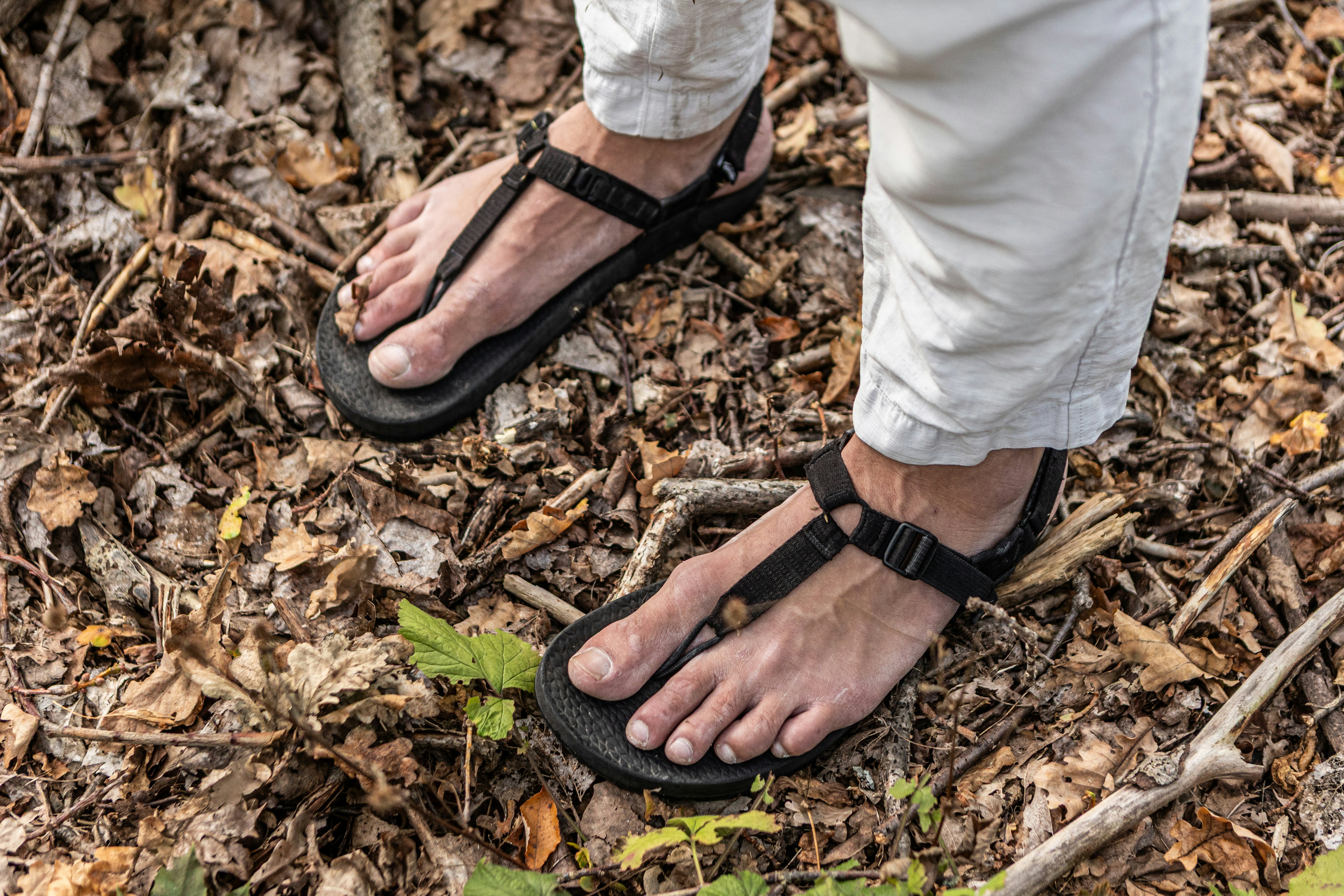 A person wearing black thong sandals and white trousers standing on dry autumn leaves