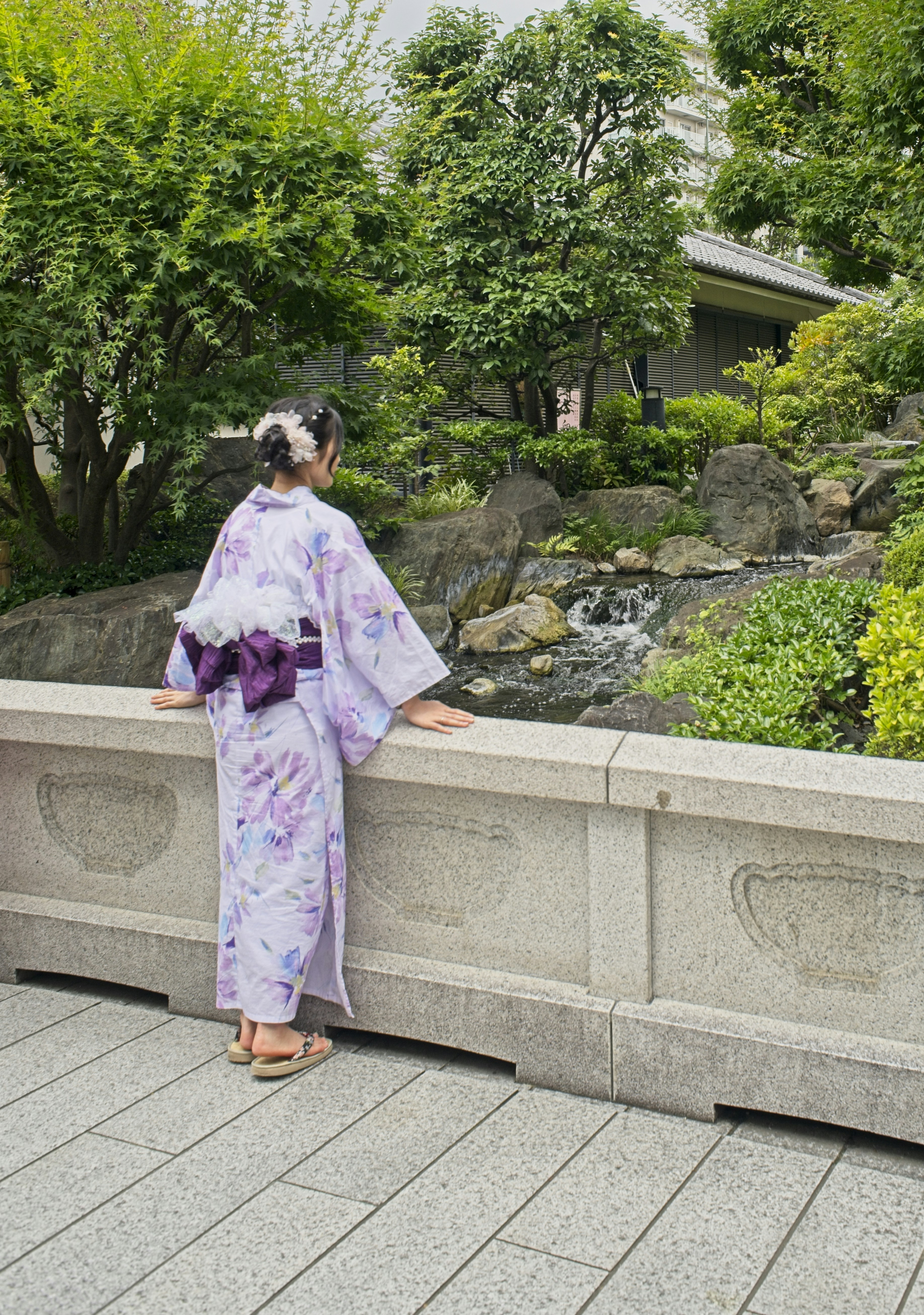 A person in a floral yukata leaning on a stone bridge by a stream
