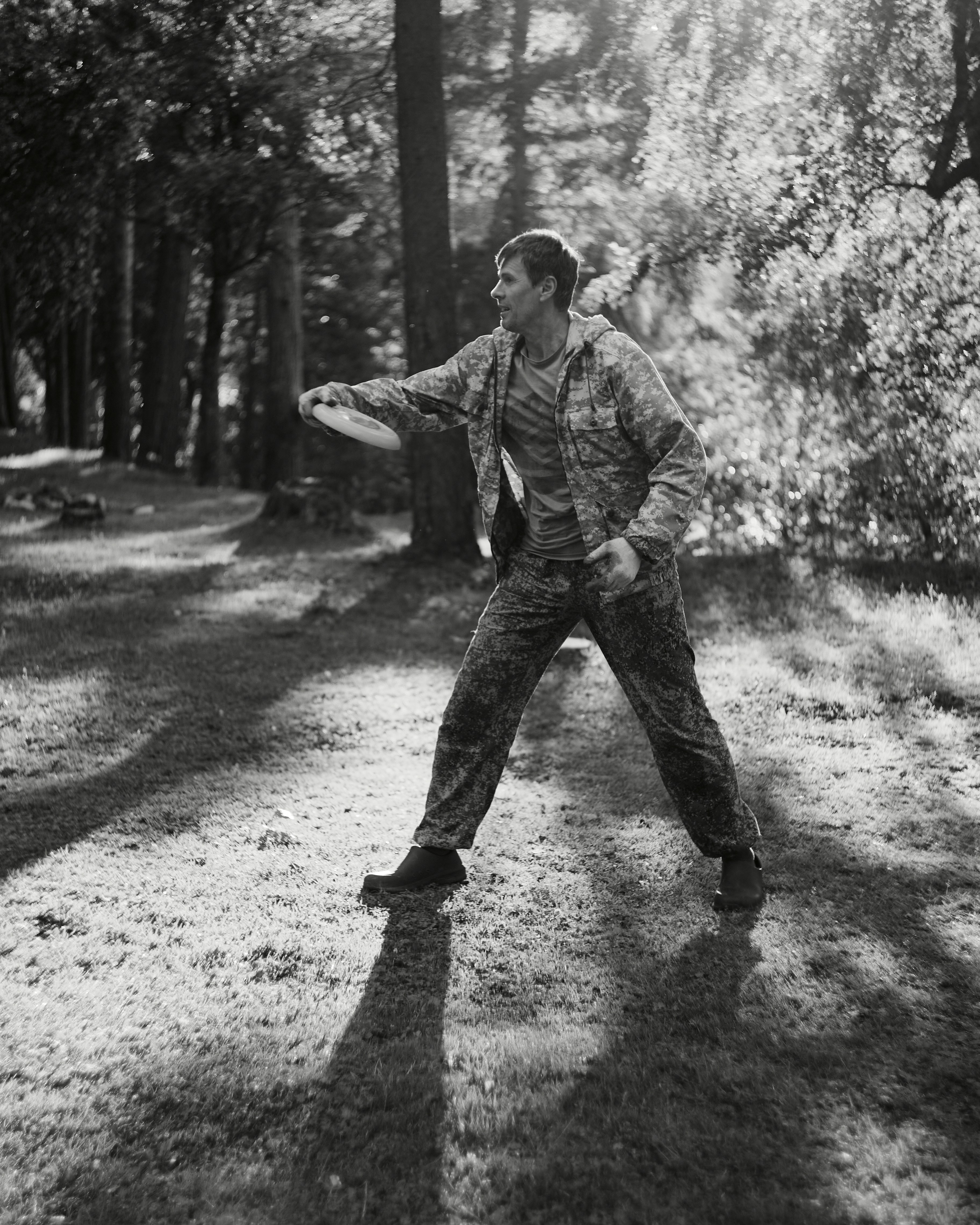 A man in camouflage clothing throwing a frisbee in a sunlit forest
