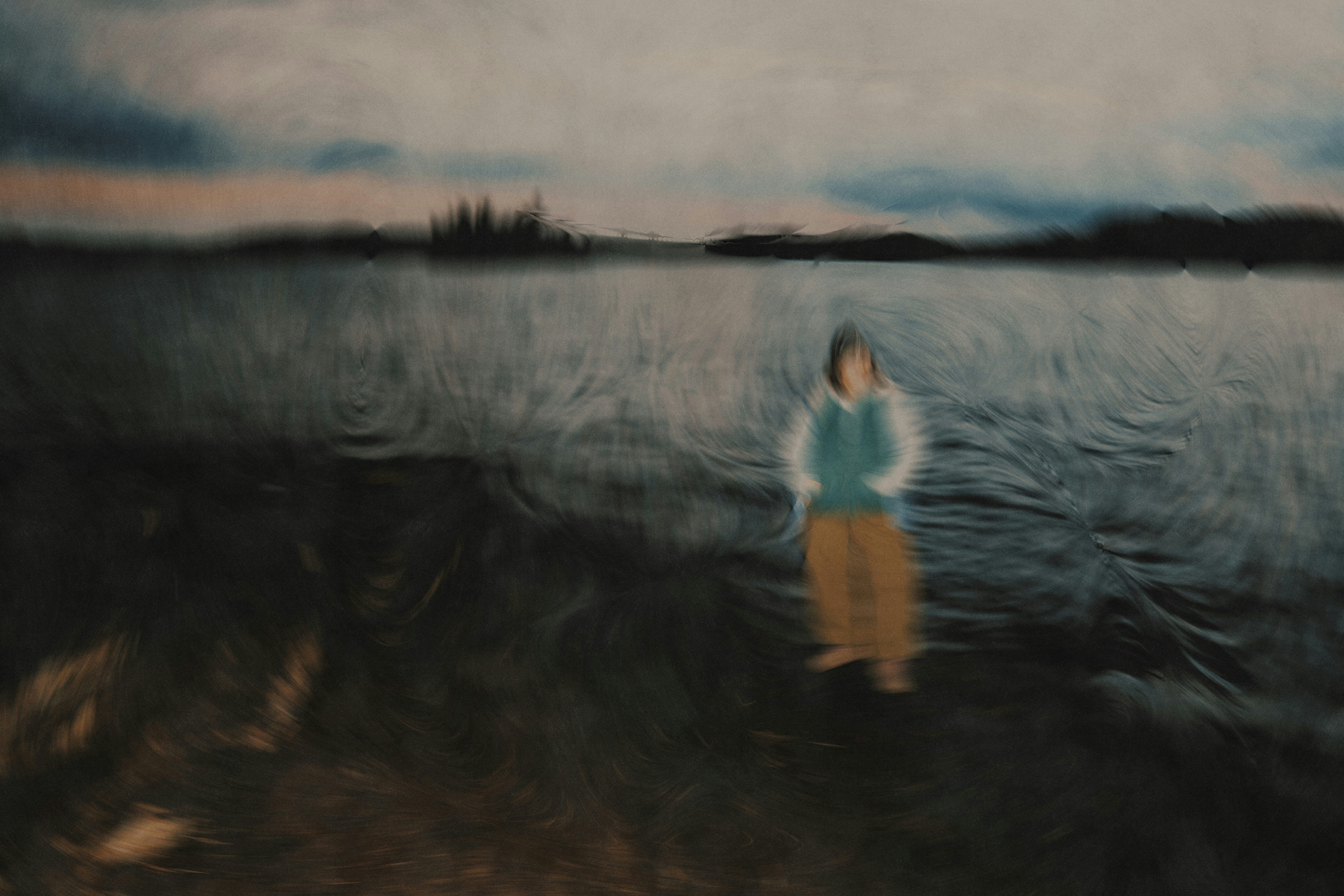 A blurred person in a teal top and brown pants by a dark lake