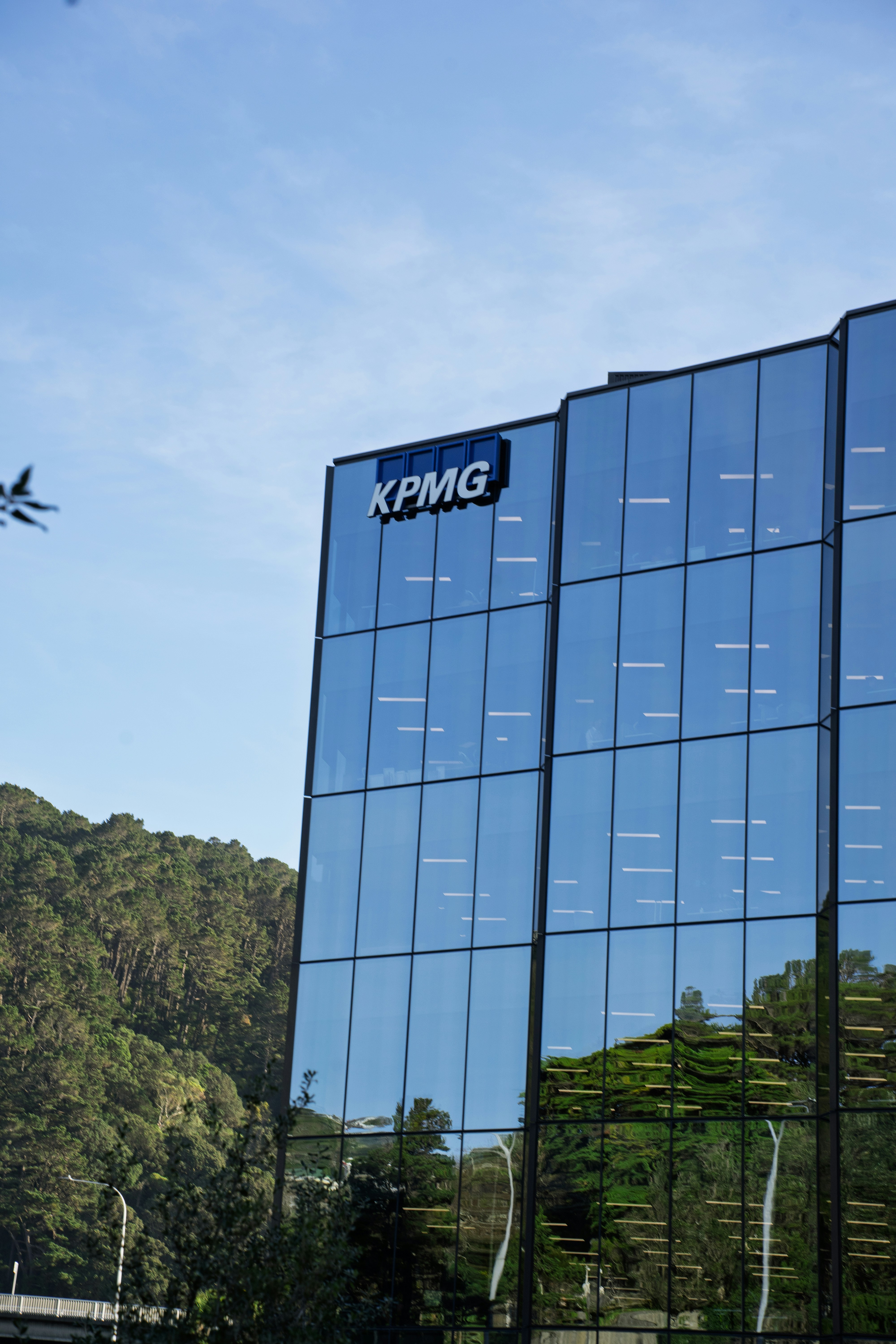 Gedung kantor kaca modern dengan logo KPMG di lereng bukit yang dipenuhi hutan