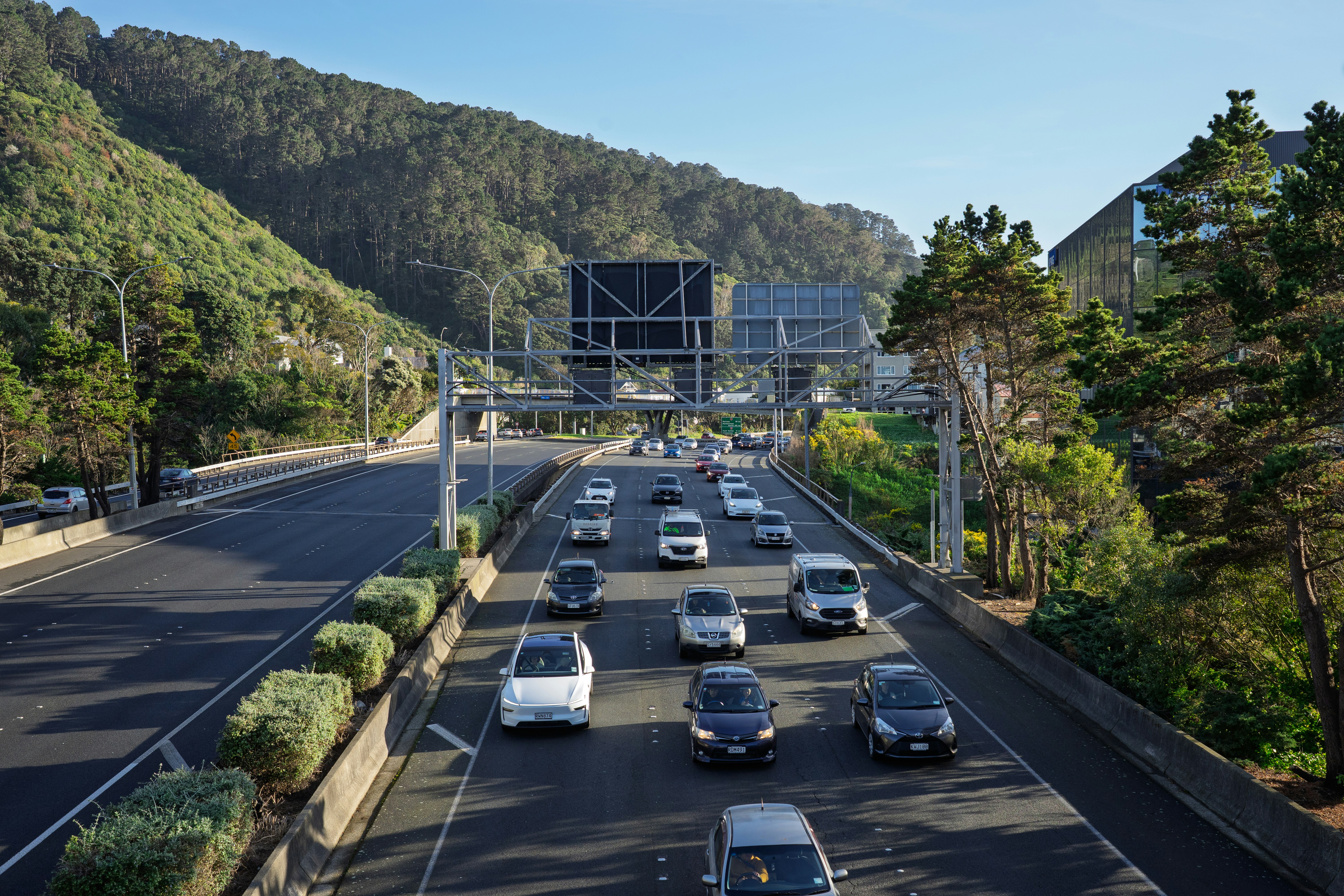 Cars driving on a multi-lane highway beside a forested hillside under a clear sky
