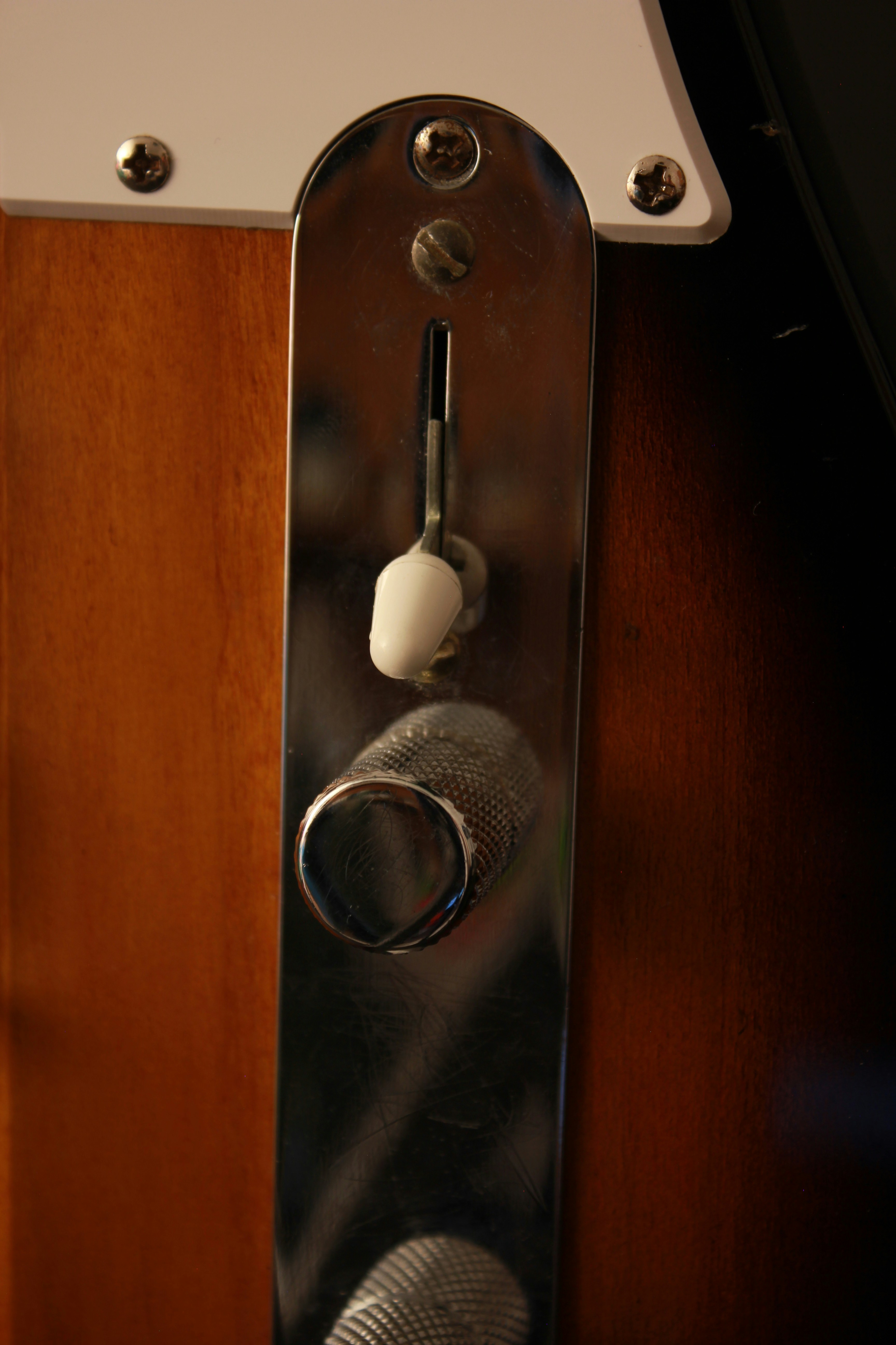 Una placa de control cromada con un interruptor blanco y un pomo moleteado en una guitarra de madera