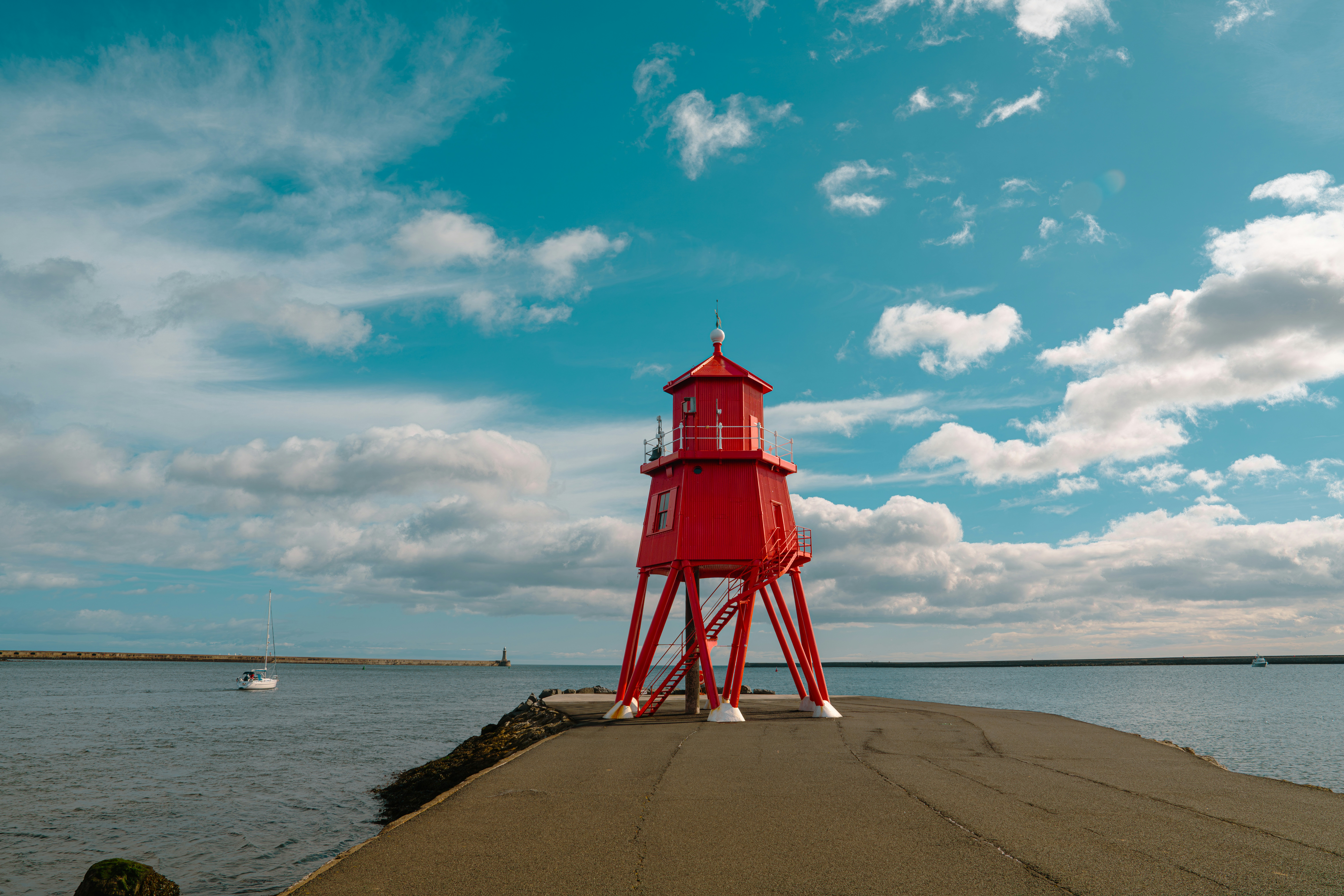 A bright red lighthouse on a concrete pier under a blue sky with clouds