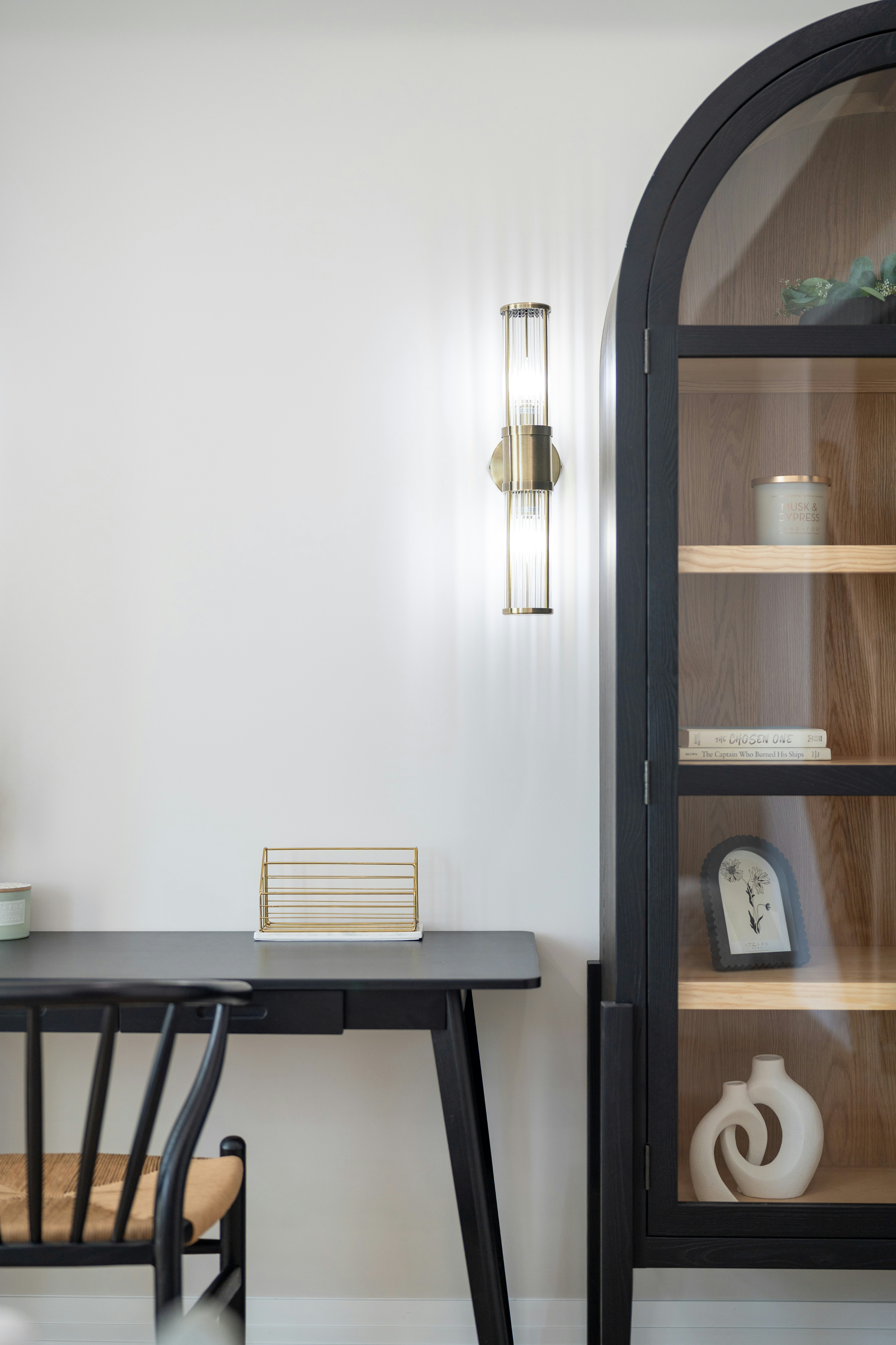 A black desk and chair beside a tall arched cabinet with glass doors