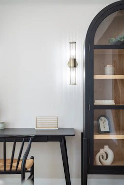 A black desk and chair beside a tall arched cabinet with glass doors