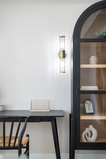 A black desk and chair beside a tall arched cabinet with glass doors