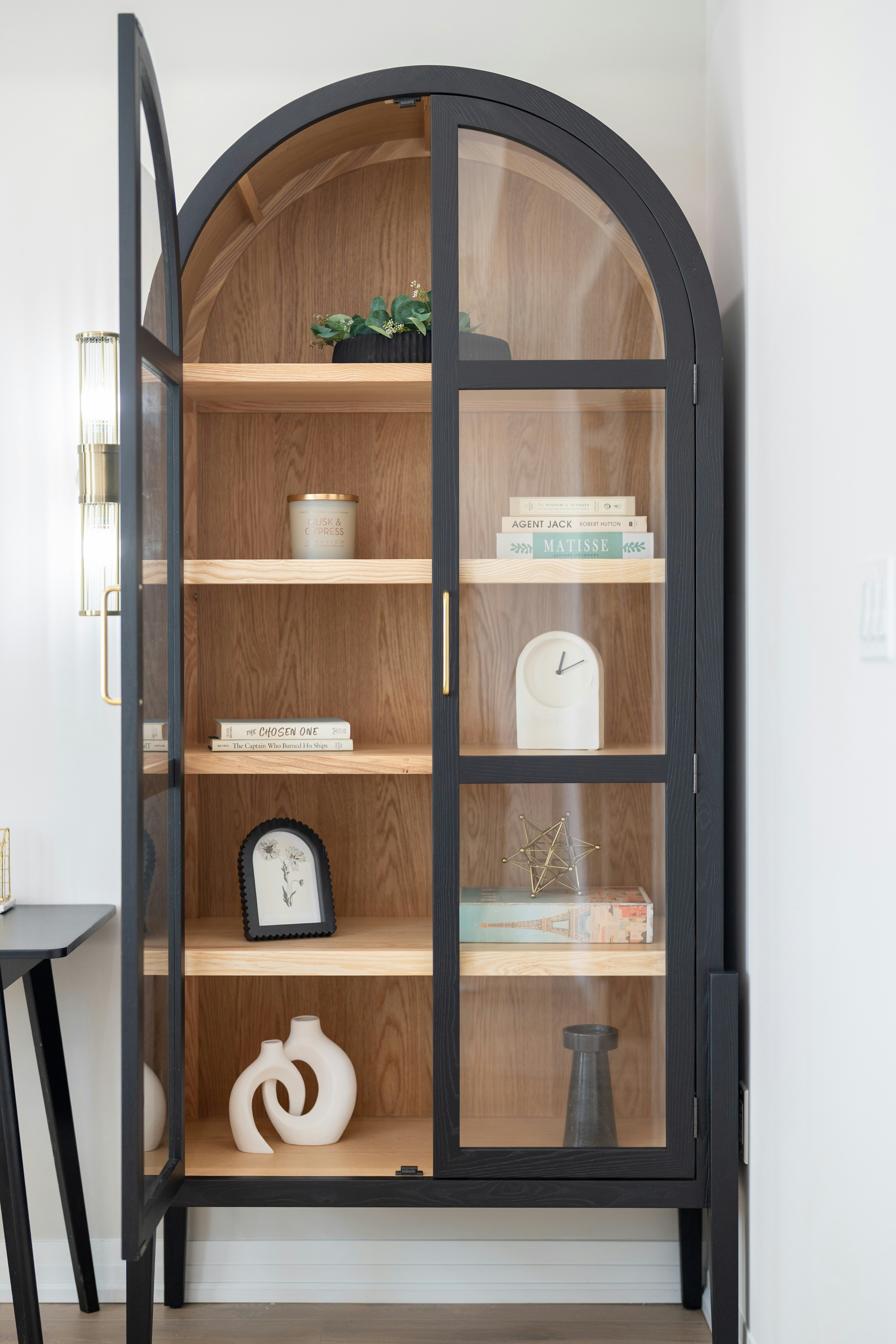 An arched black cabinet with glass doors displaying books and decorative objects
