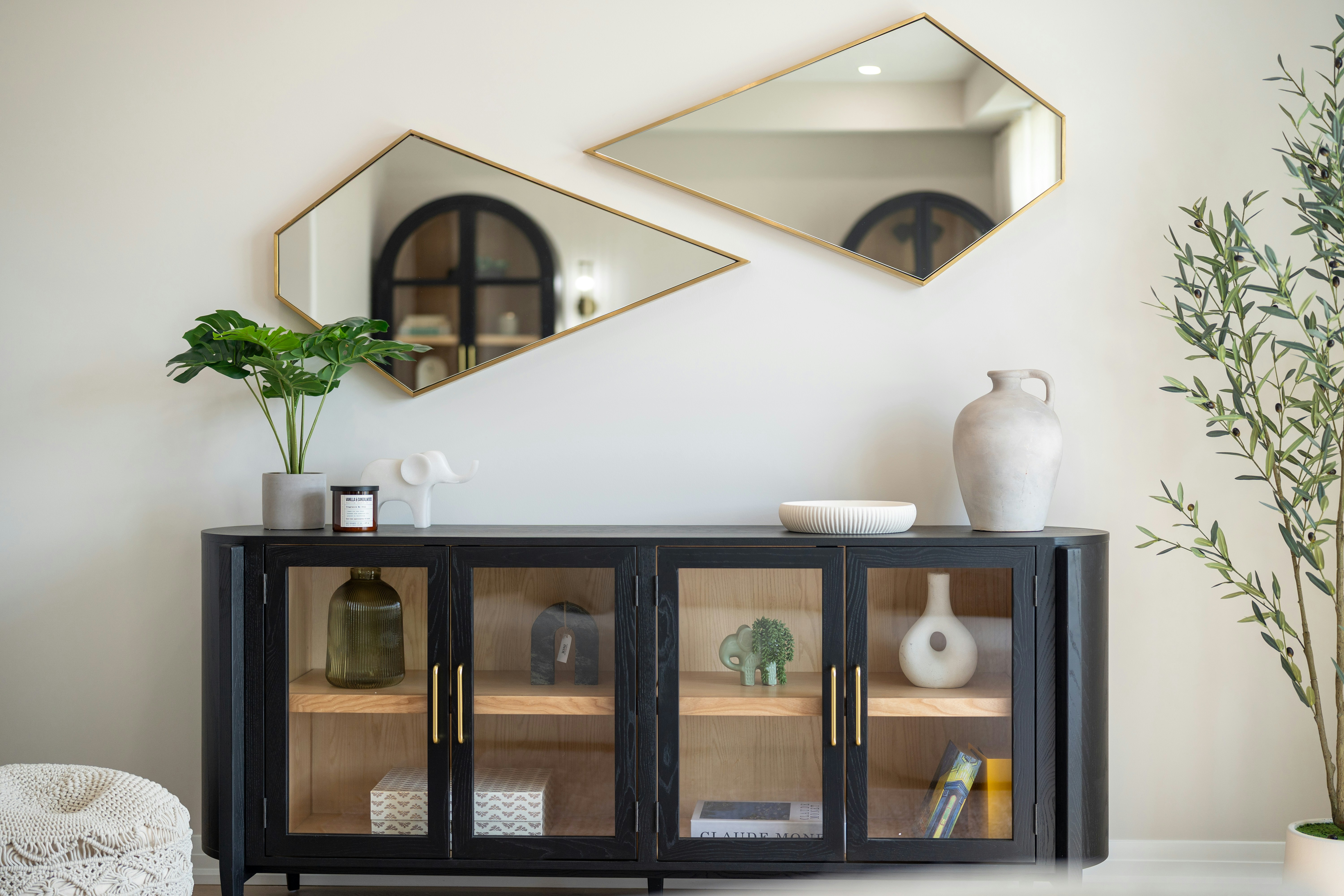 A black wooden sideboard with glass doors beneath two geometric gold-framed mirrors