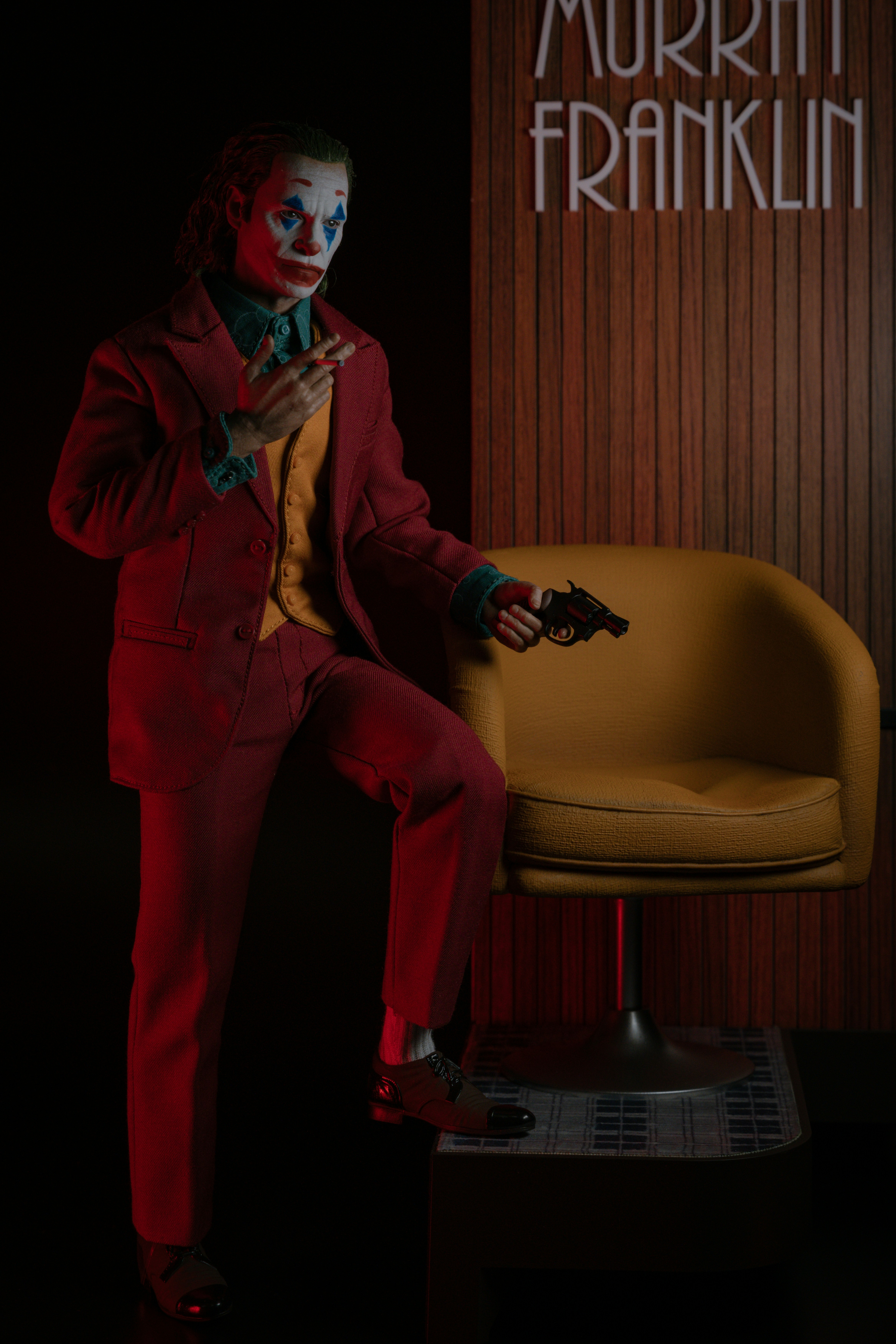 A person in clown makeup and a red suit holding a cigarette and handgun