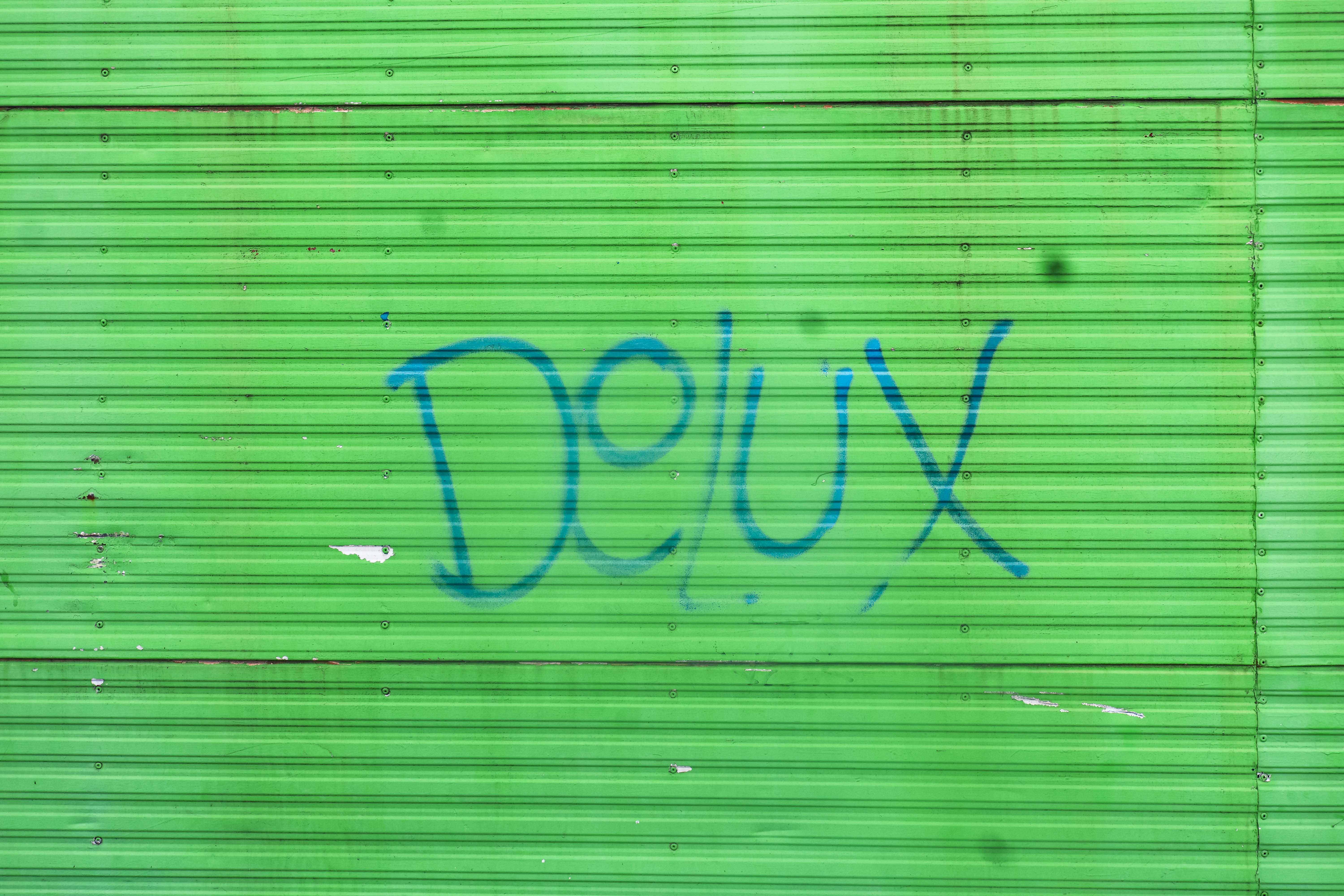 밝은 녹색 골판지 금속 벽에 "Delux"라고 적힌 파란색 그래피티가 있다