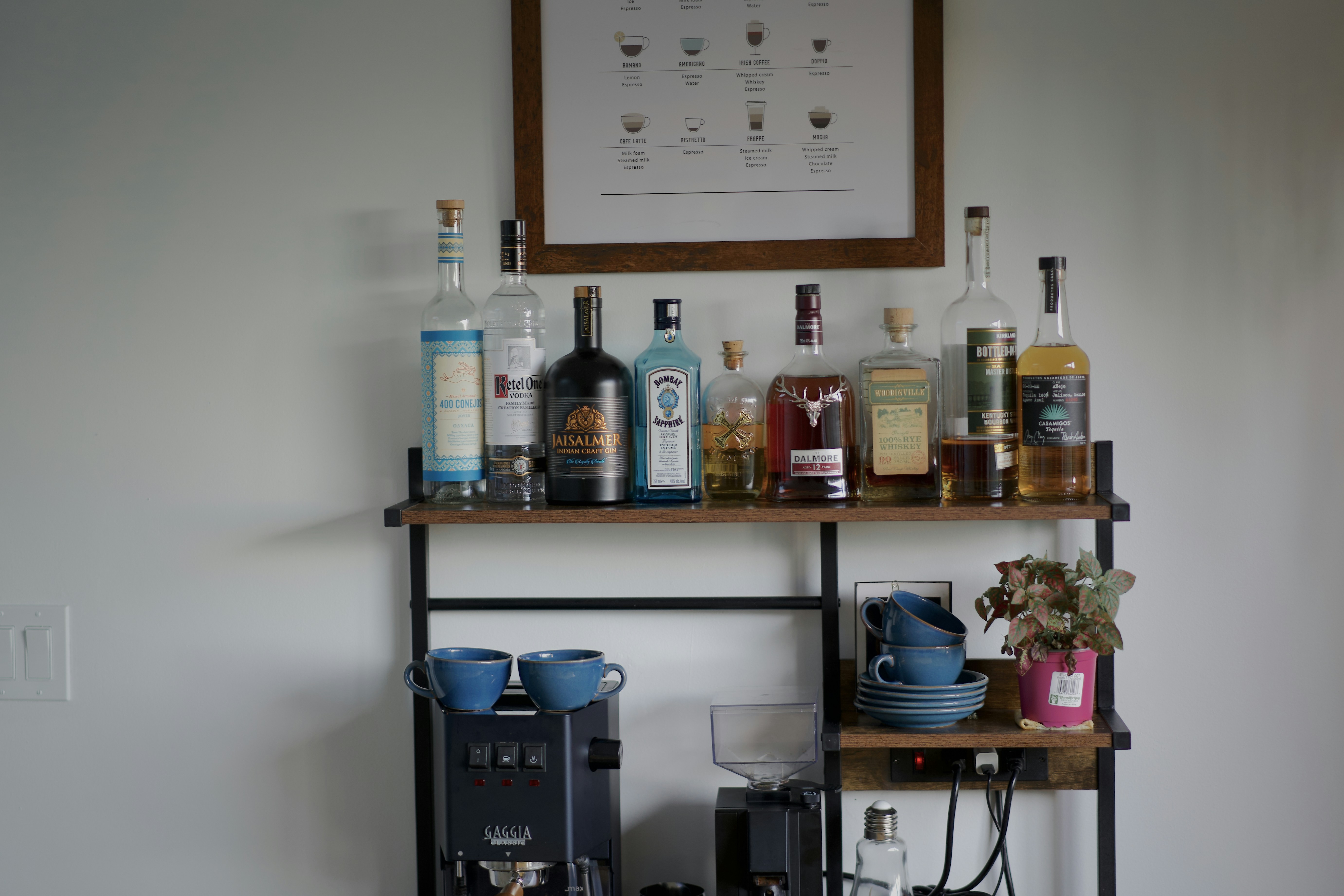 A compact home coffee station beneath a shelf of bottles.