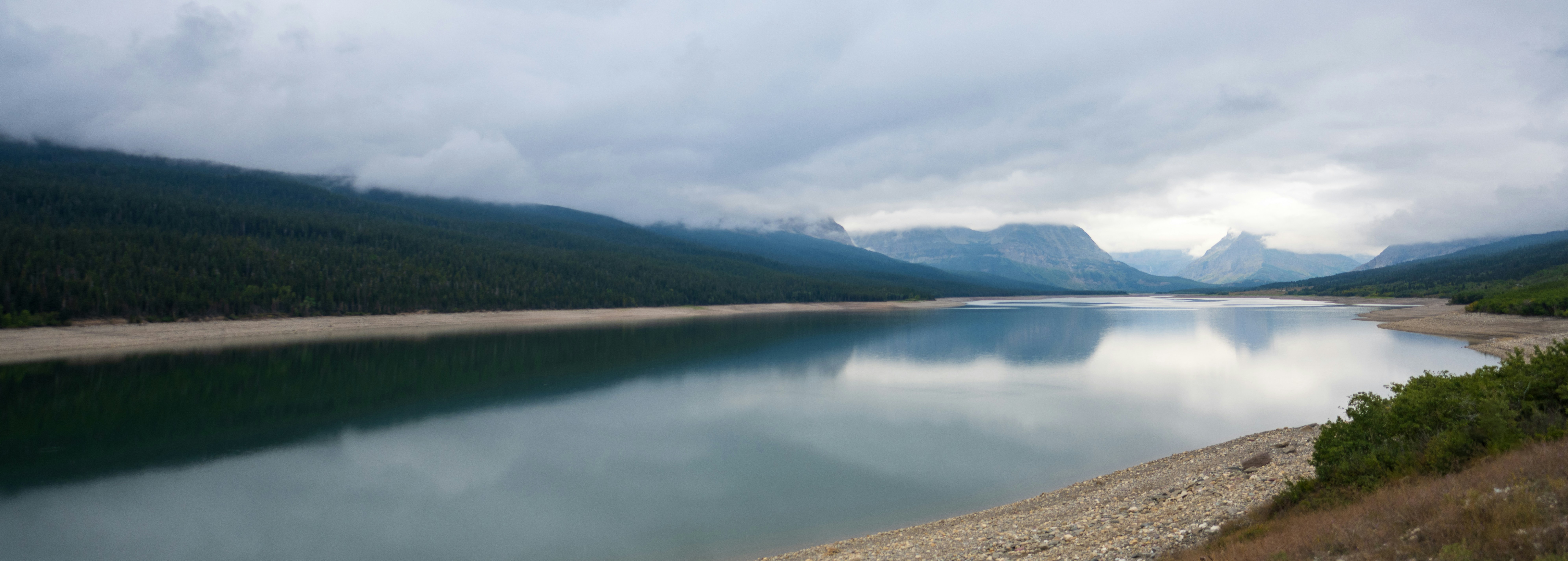 A calm lake reflecting cloudy mountains and a dense pine forest