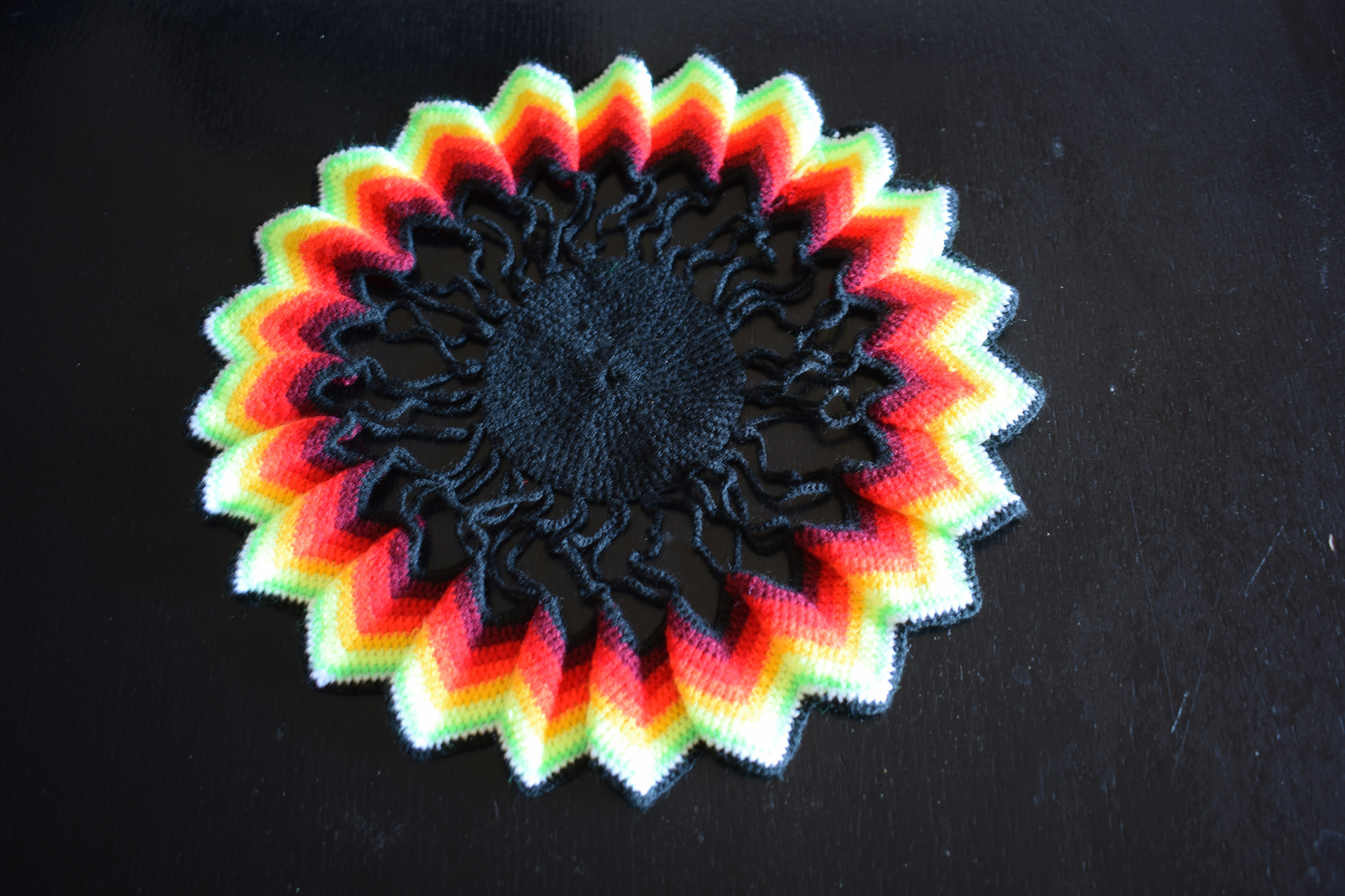 A circular crocheted doily with a black center and rainbow colored zigzag edges