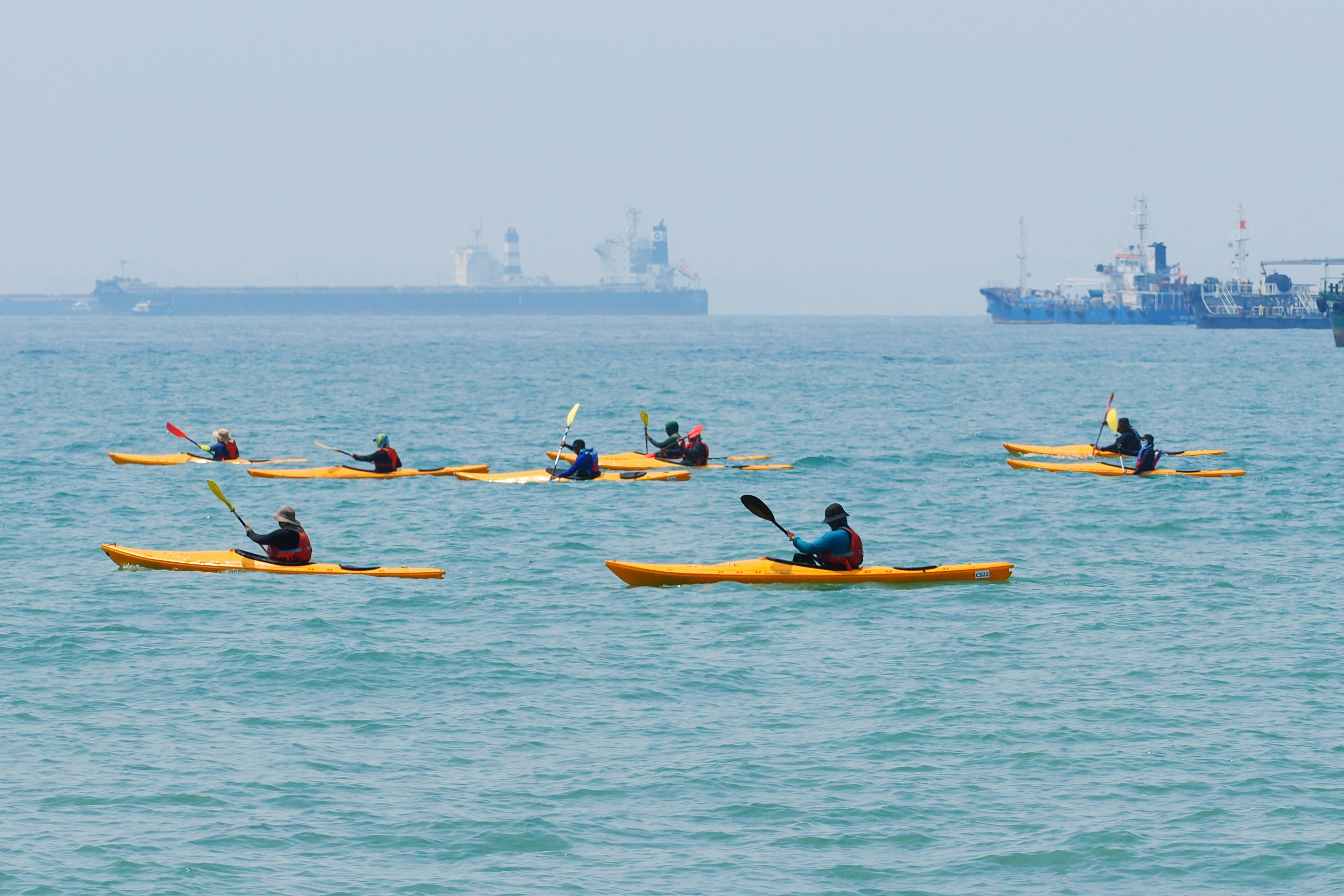 A group of people paddling yellow kayaks on the open sea near large ships