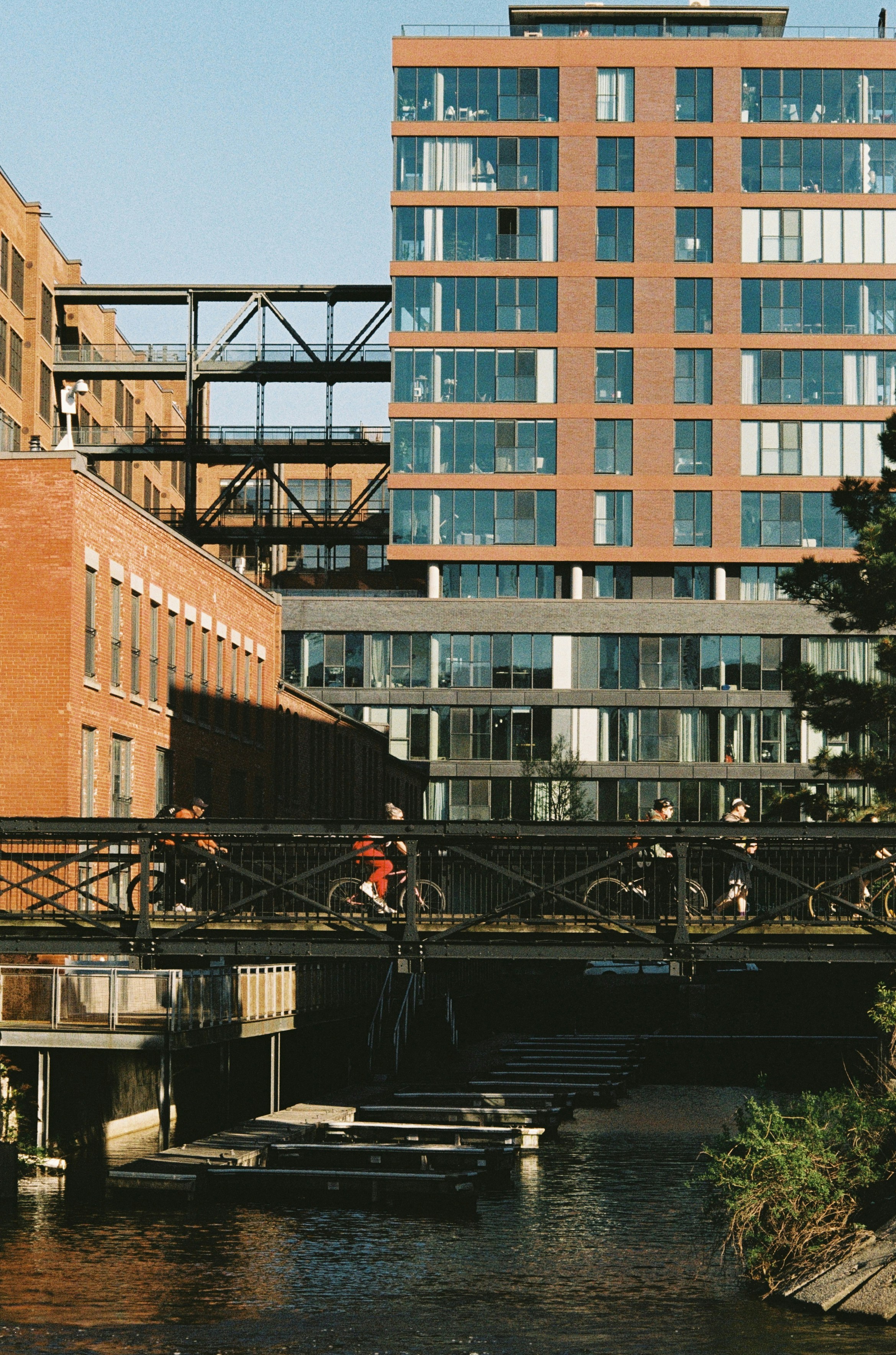 People cycling across a metal bridge over a canal near brick buildings