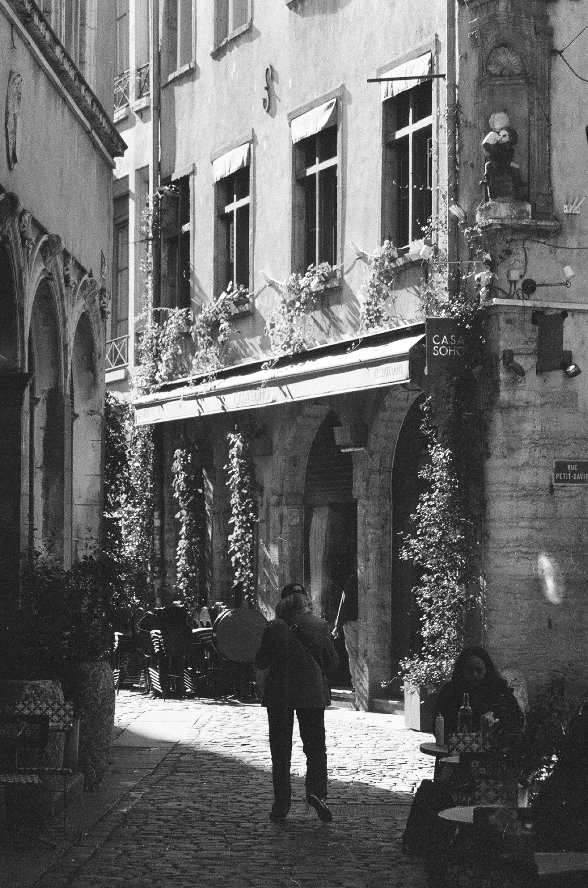 A person walking down a narrow cobblestone street past a restaurant with climbing vines
