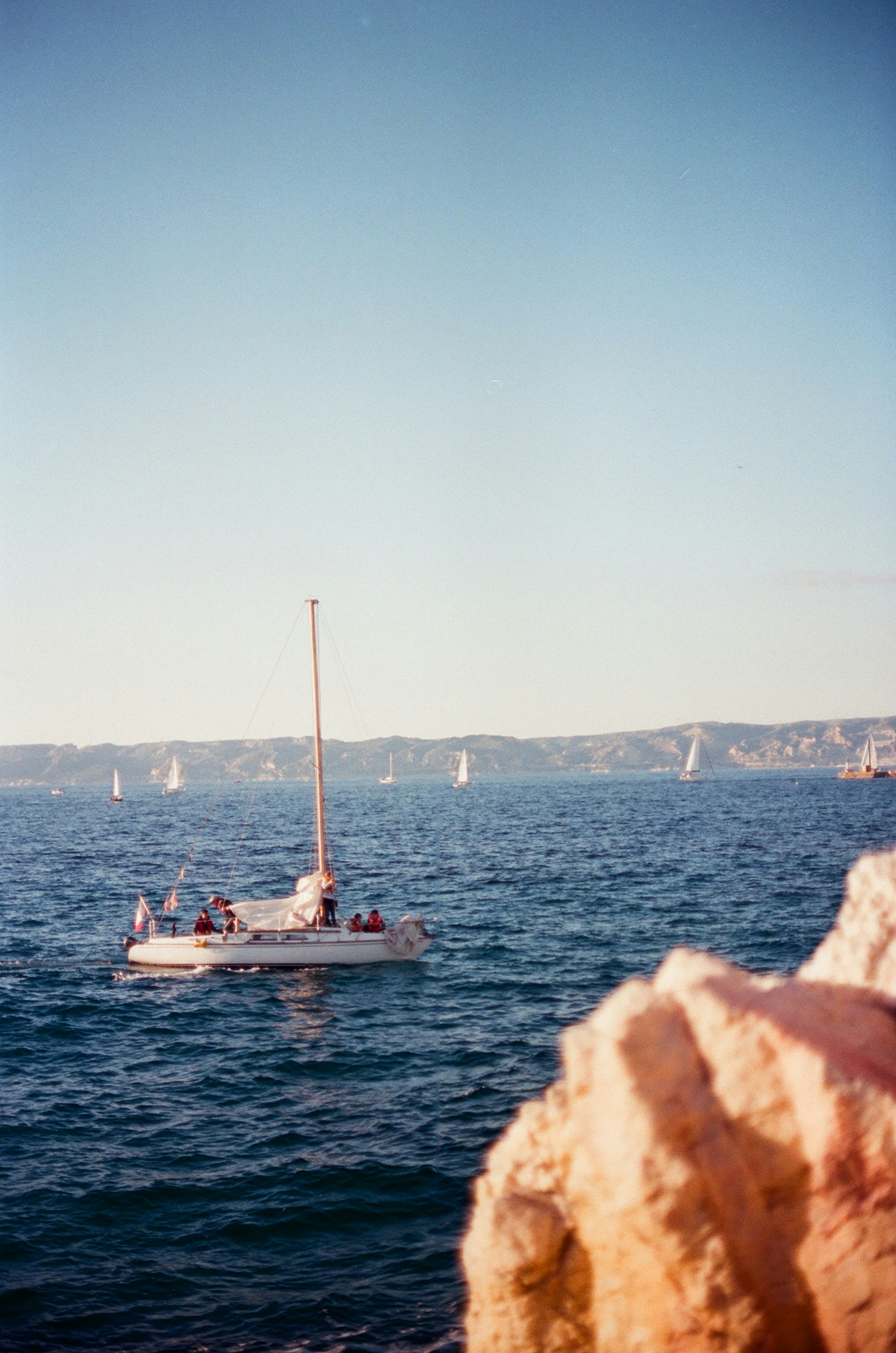 A sailboat with people on deck on blue water near a rocky coastline