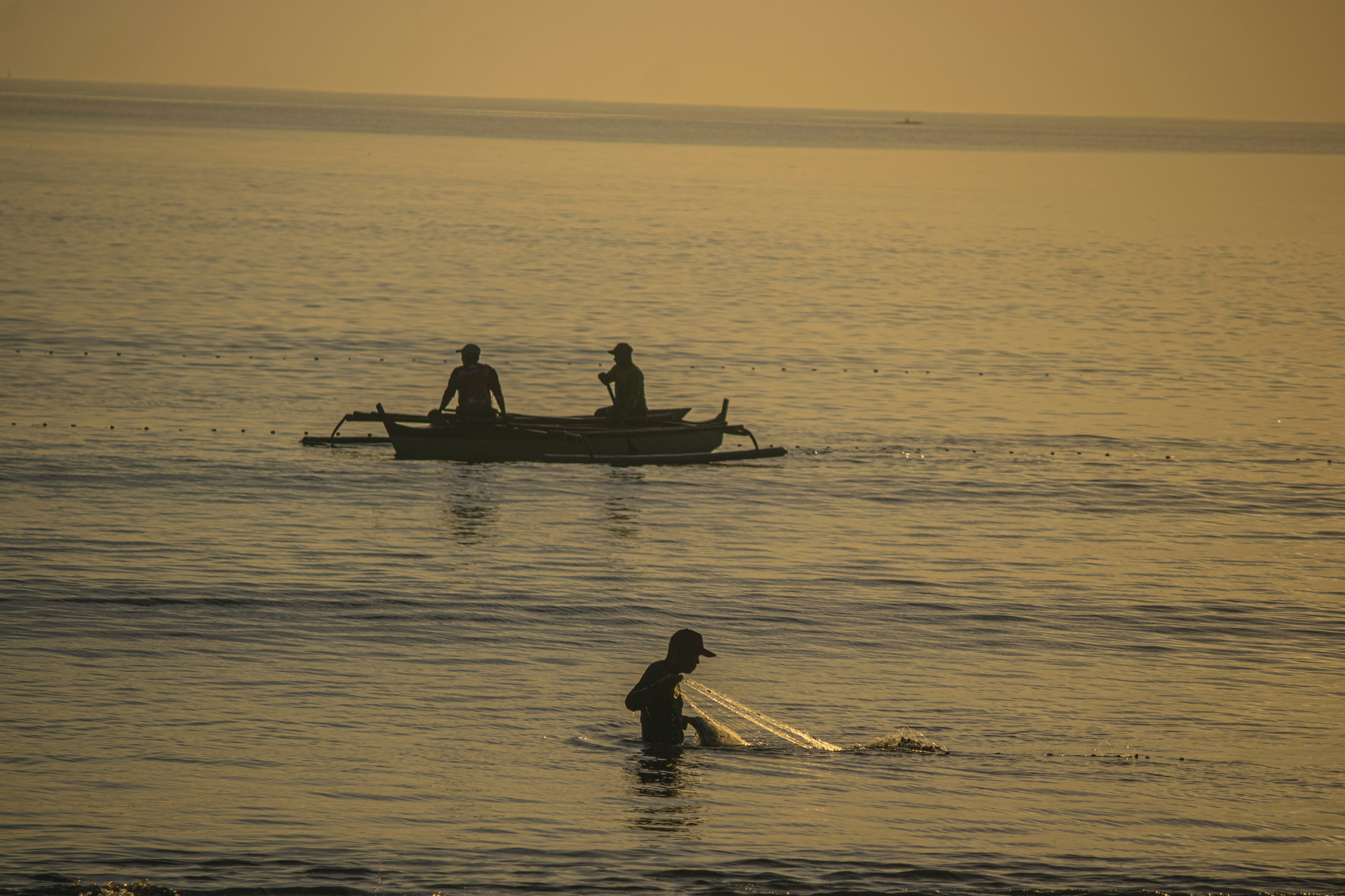 A person casting a fishing net in shallow water near a boat with two people