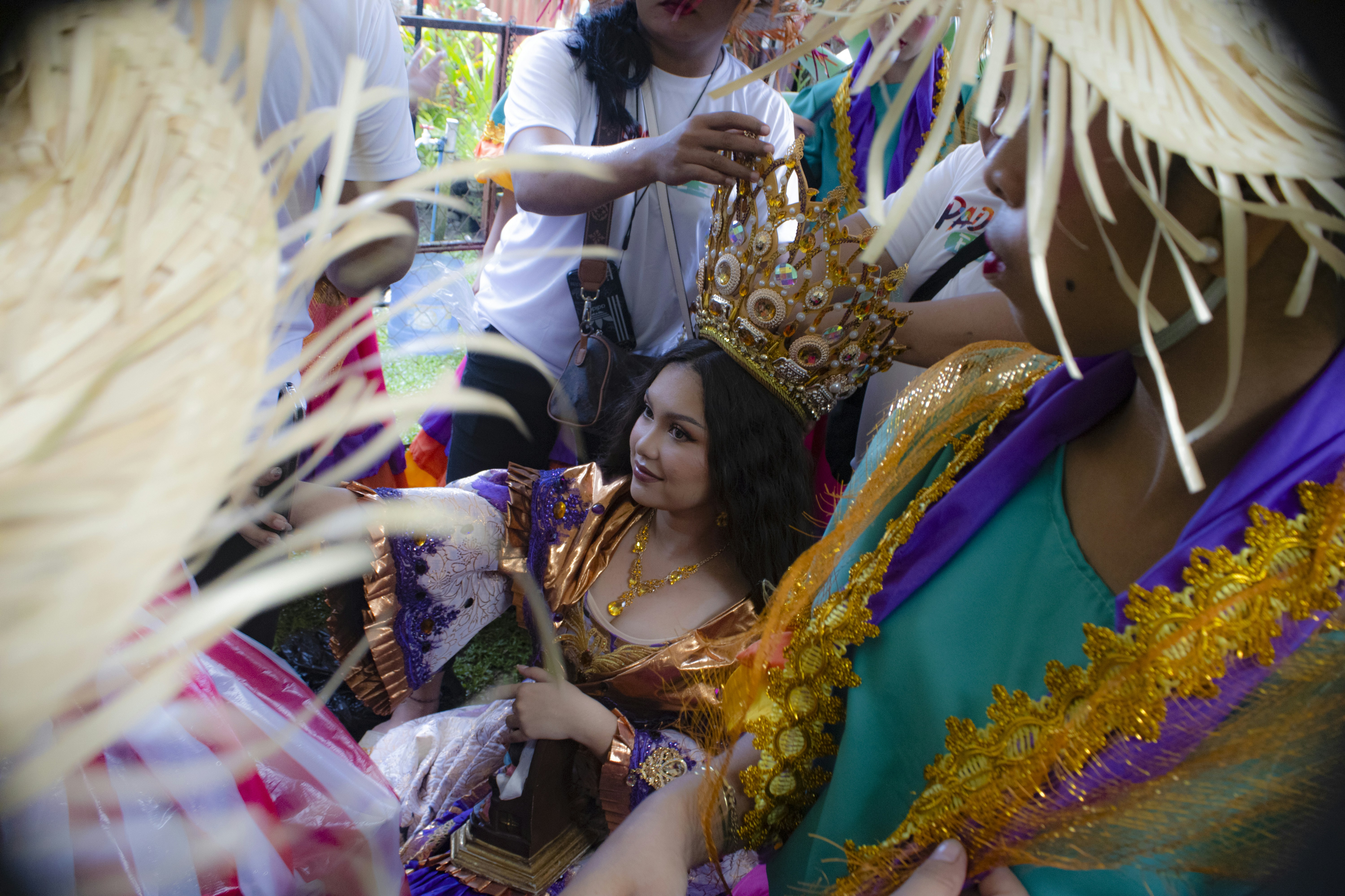 A woman in a metallic dress wearing a large crown while people assist her