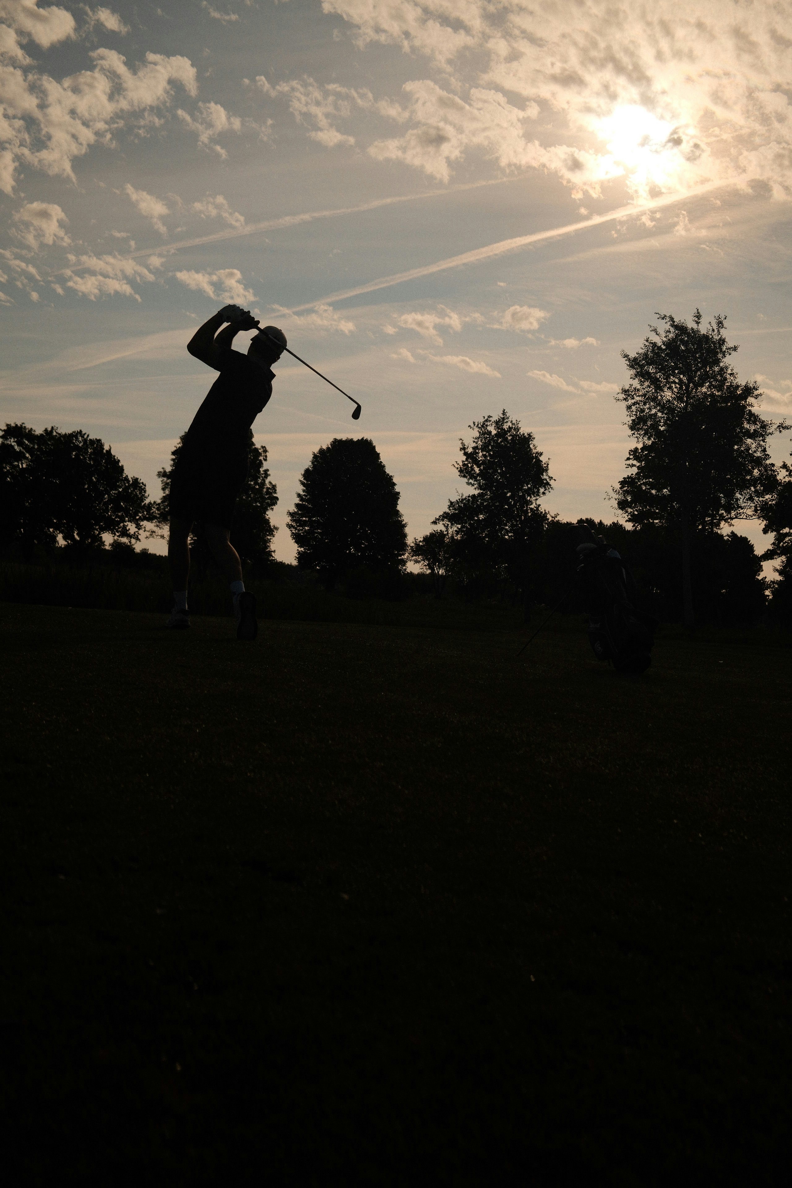 A golfer in silhouette swinging a club against a bright sky with clouds