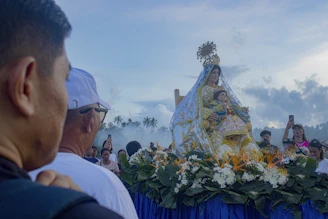 A crowd of people watching a religious statue on a flower-covered float outdoors