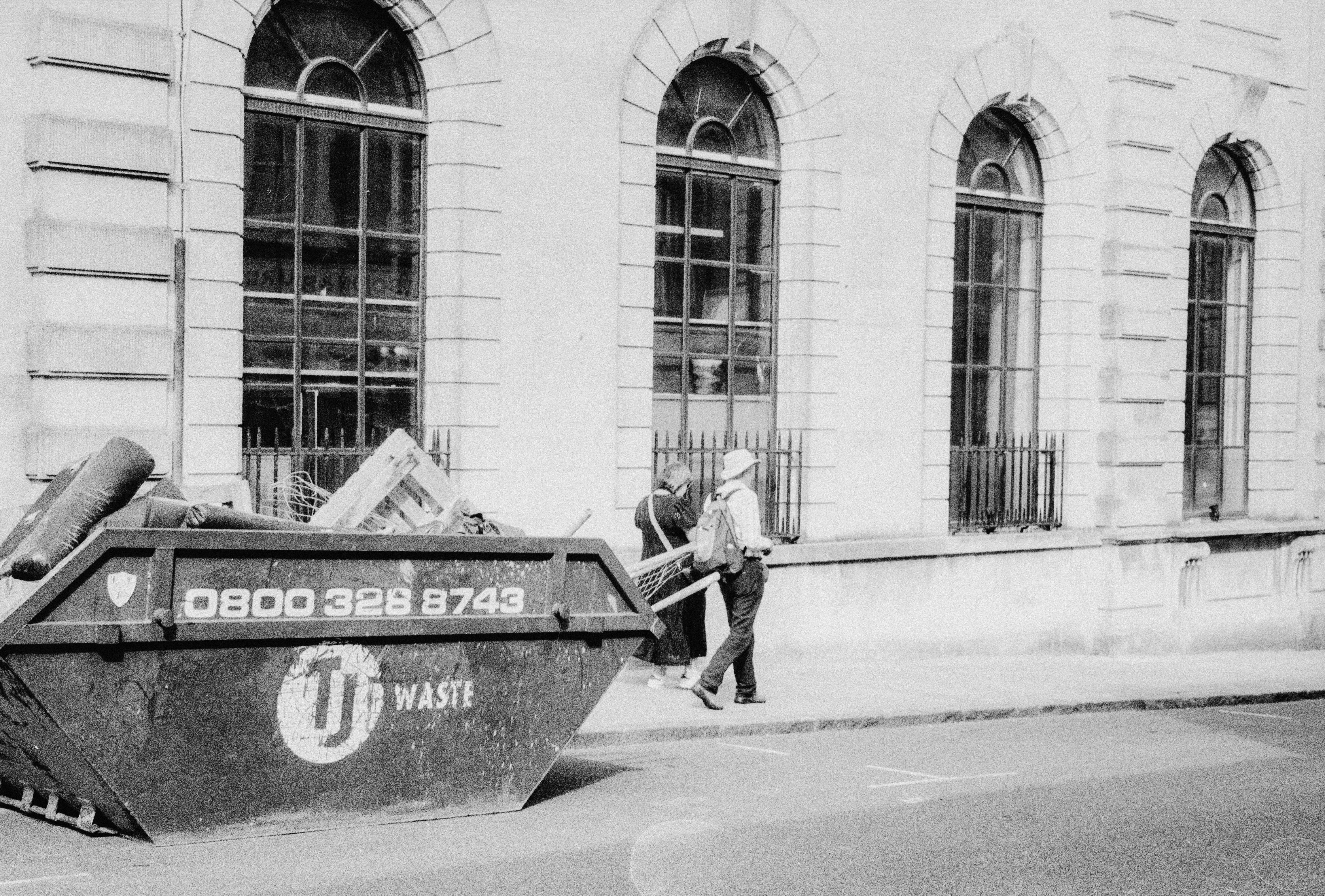 Two people walking past a large metal waste skip on a city sidewalk
